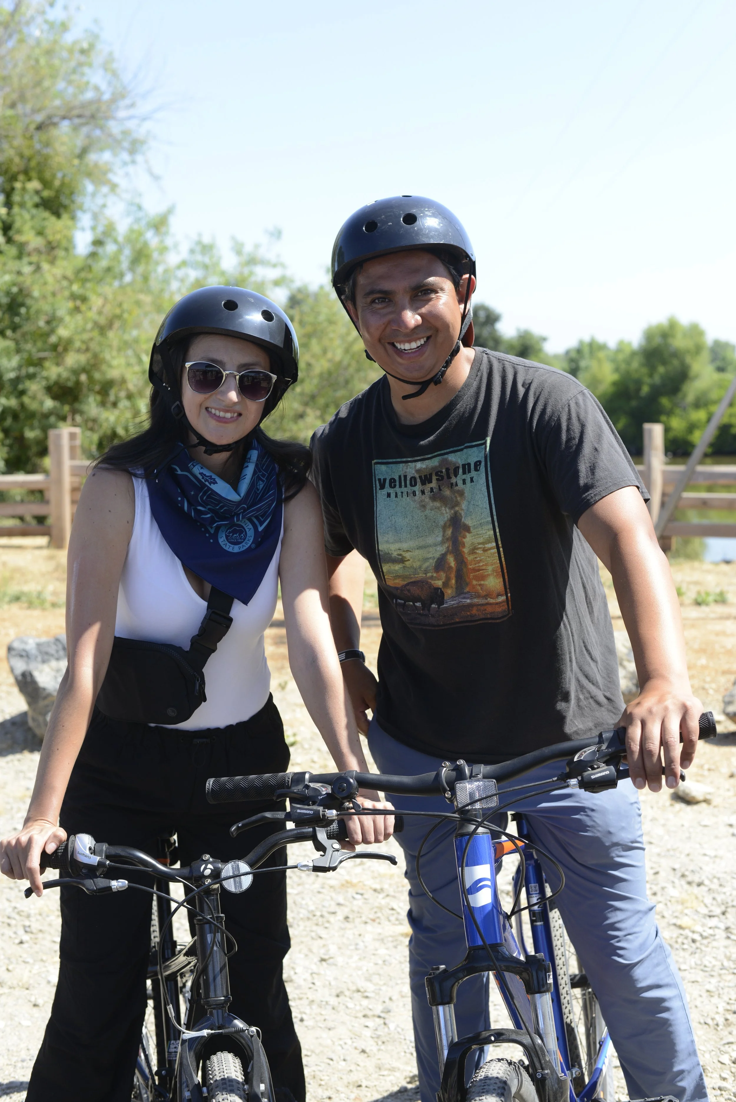 A man and woman wearing helmets and sunglasses, smiling, standing with bikes outdoors on a sunny day.