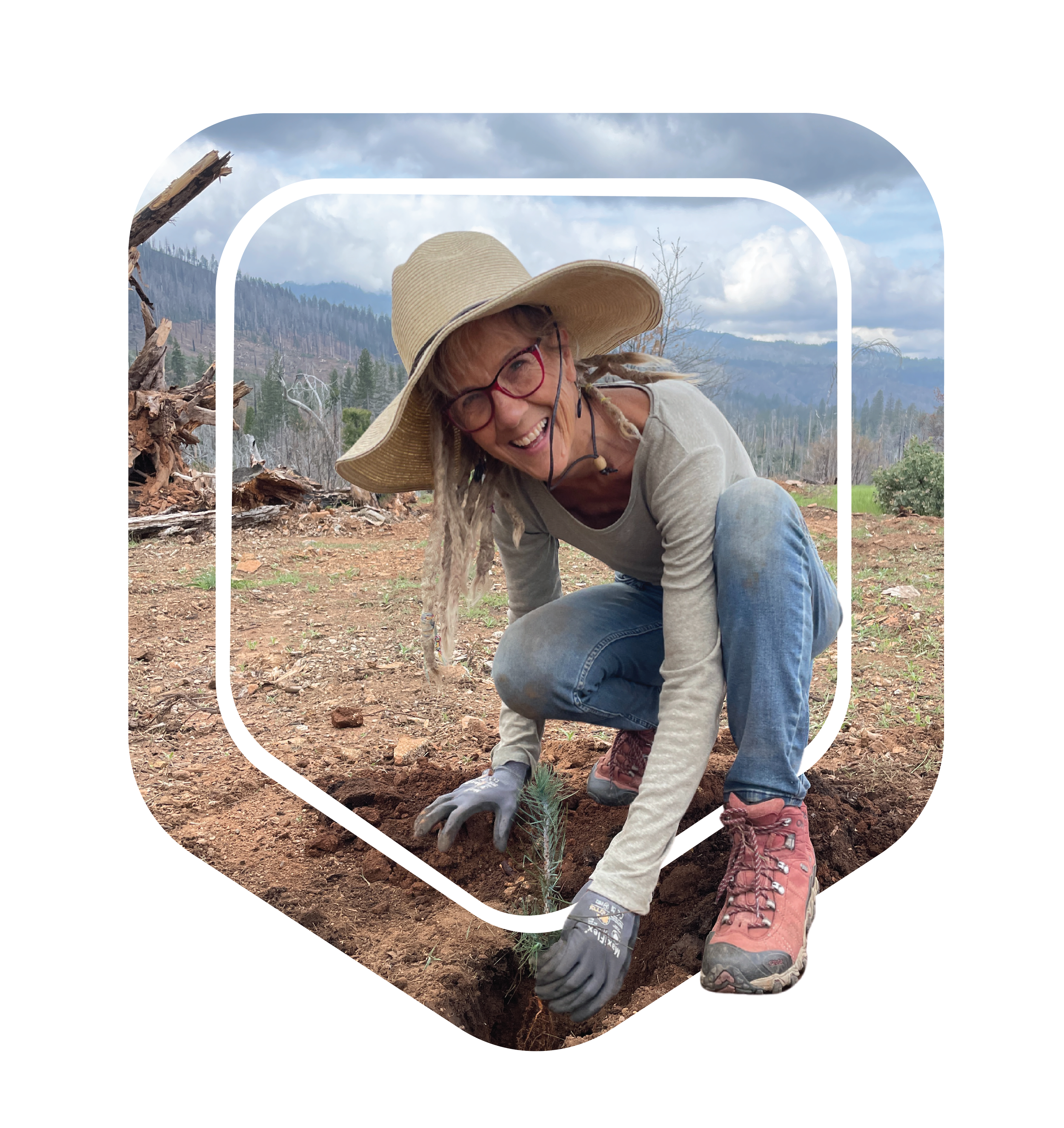 Woman planting a small tree in a dirt patch outdoors, wearing a wide-brimmed hat, glasses, gray sweater, jeans, and pink boots, with a mountainous landscape and cloudy sky in the background.