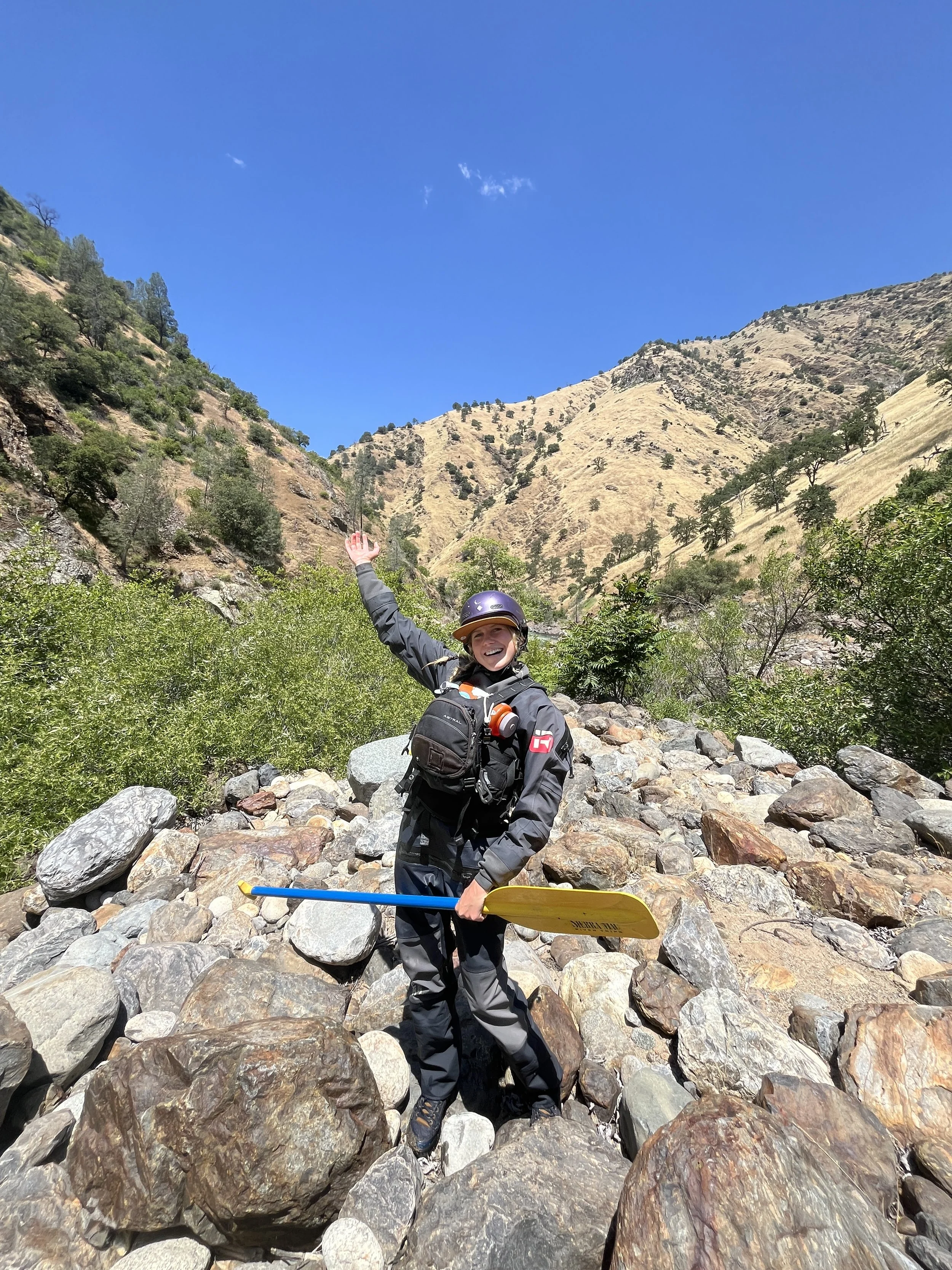 A person in outdoor gear, wearing a helmet and carrying a backpack, smiling and waving while holding a paddle in a rocky riverbed with mountainous landscape in the background.