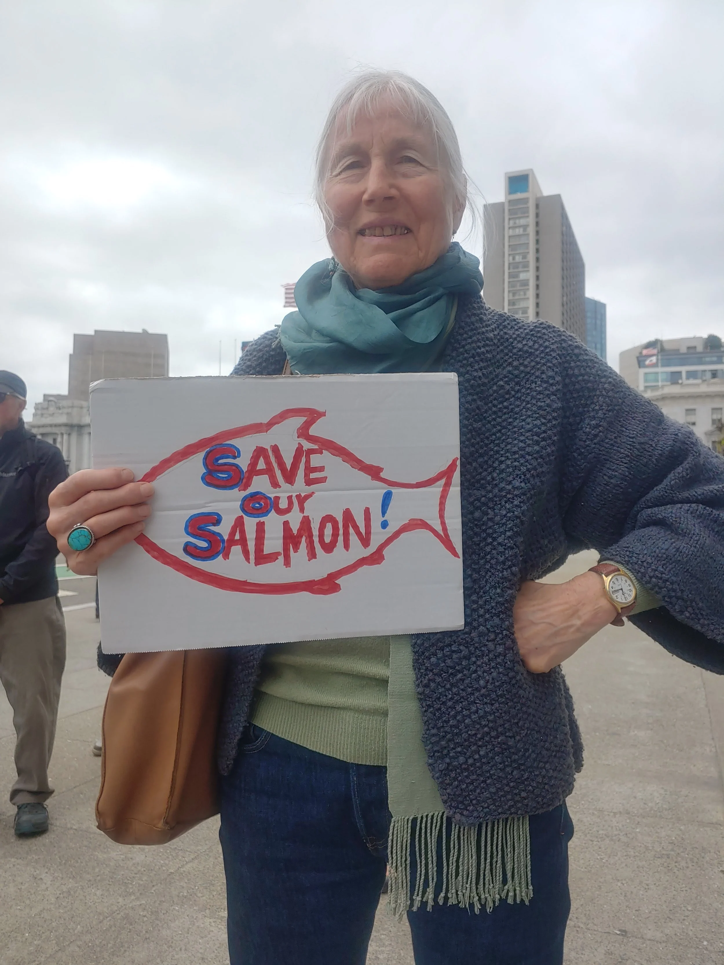 A woman holding a sign that says, 'Save Our Salmon!' at an outdoor protest in a city with tall buildings in the background.
