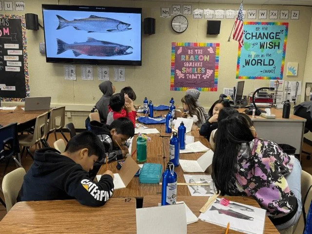 A classroom with students sitting at tables, some writing and others looking at a large screen displaying images of fish. The classroom walls are decorated with colorful posters and alphabet letters.