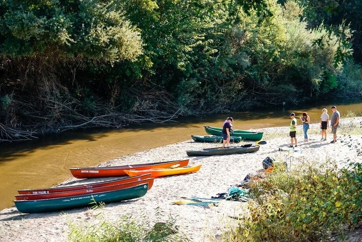 People preparing canoes and kayaks on a sandy riverbank with green trees in the background.