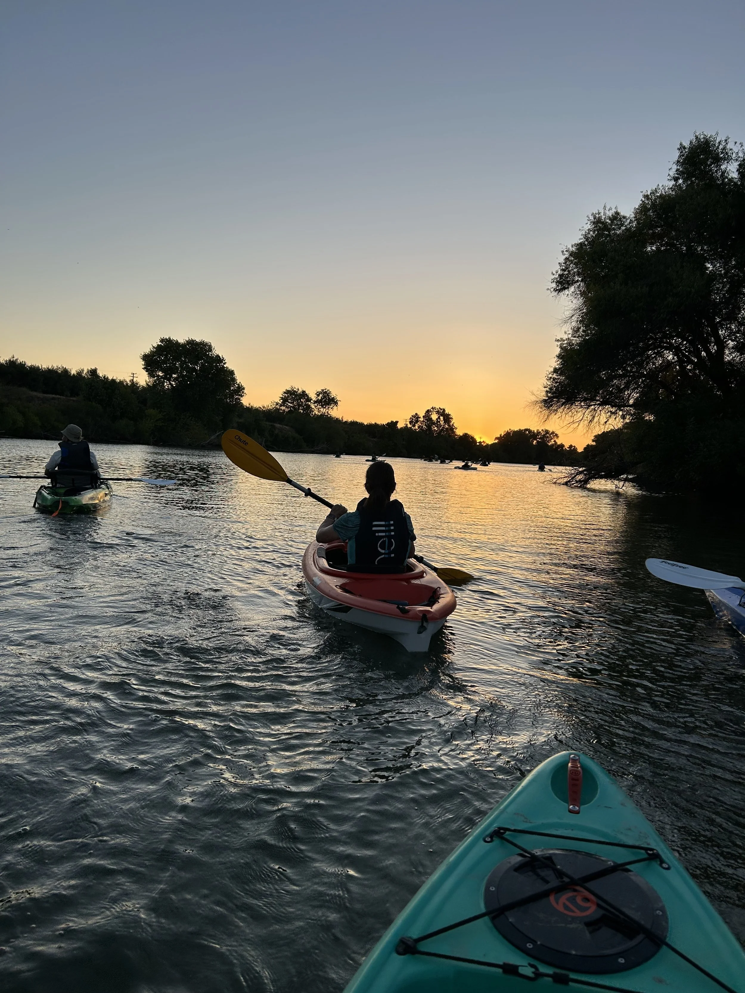 People kayaking on a river at sunset, with trees along the water's edge and small boats in the distance.