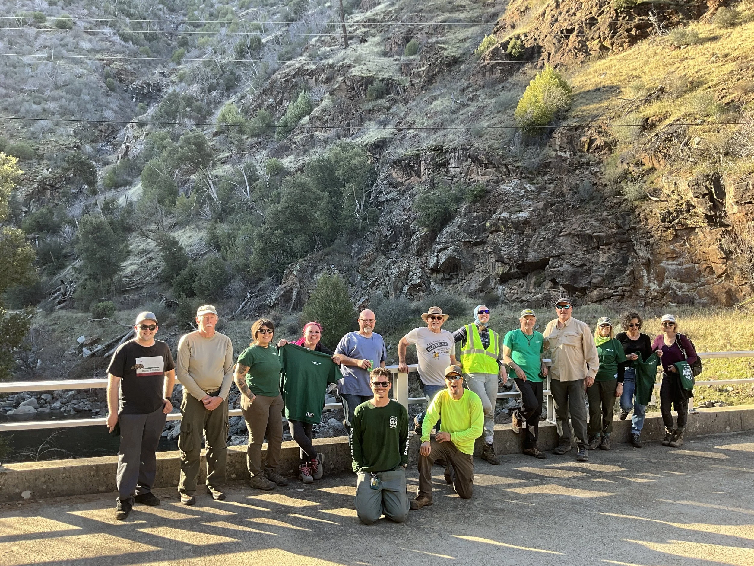 Group of people standing outdoors on a bridge with a rocky hillside and trees in the background.