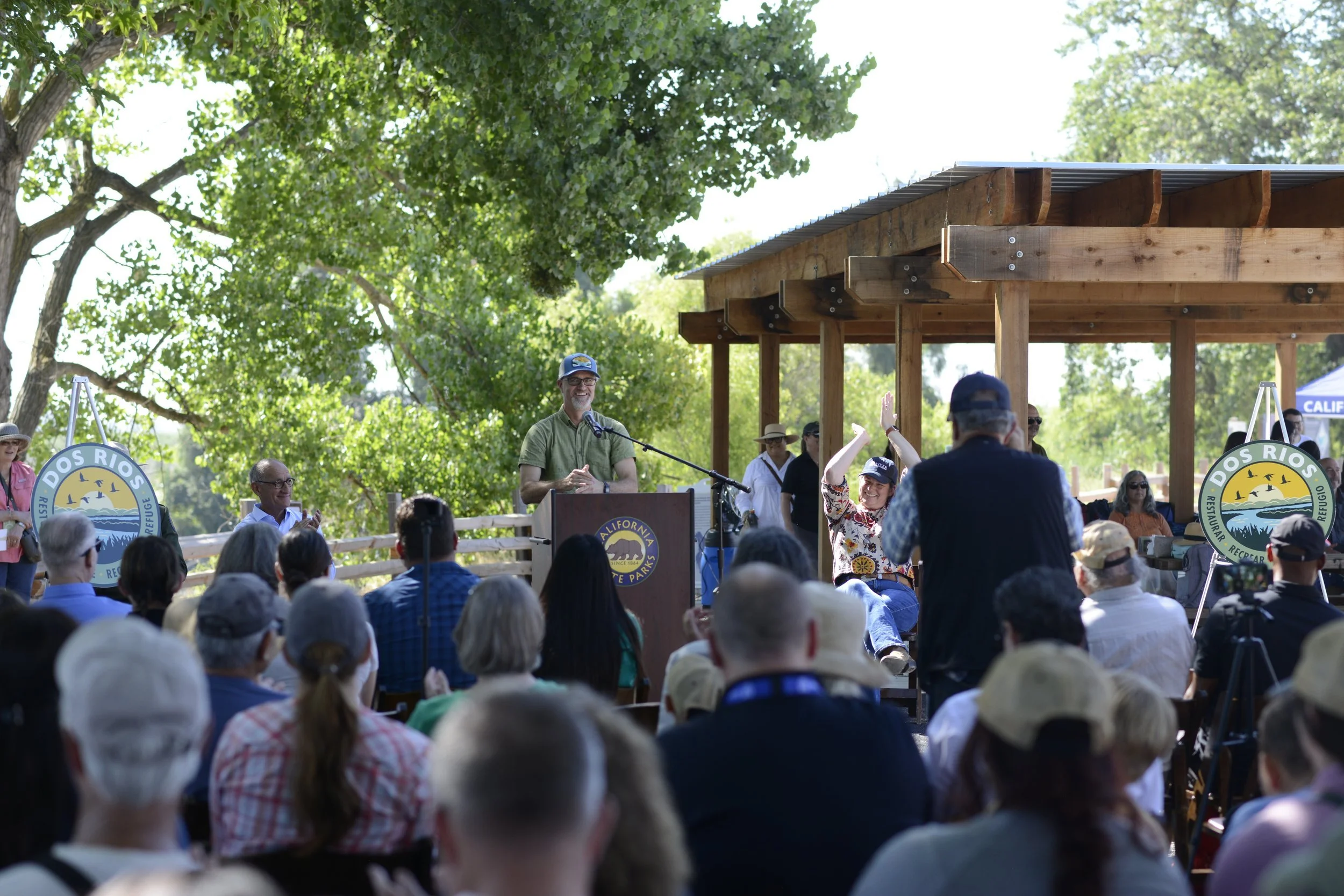 A man at a podium speaking to an outdoor crowd during an event, with people seated and standing, some clapping and raising their hands, surrounded by trees and banners for Dos Rios and California State Parks.