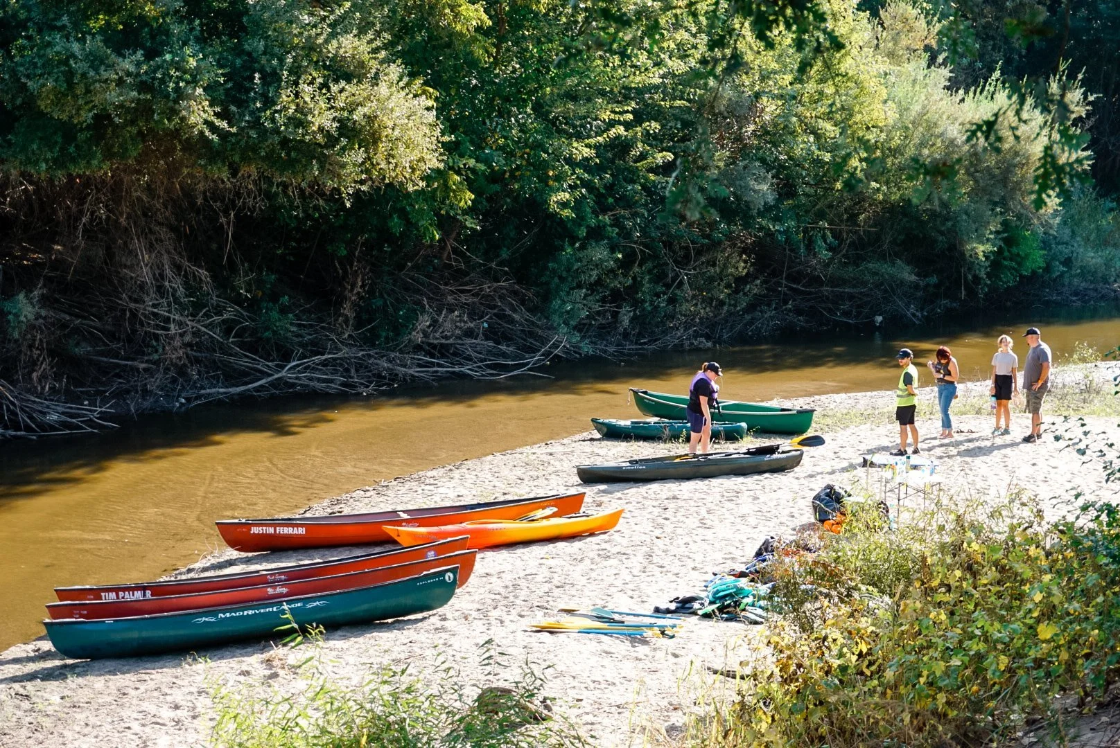 Group of people preparing canoes and kayaks on a sandy riverside, with dense green trees in the background.