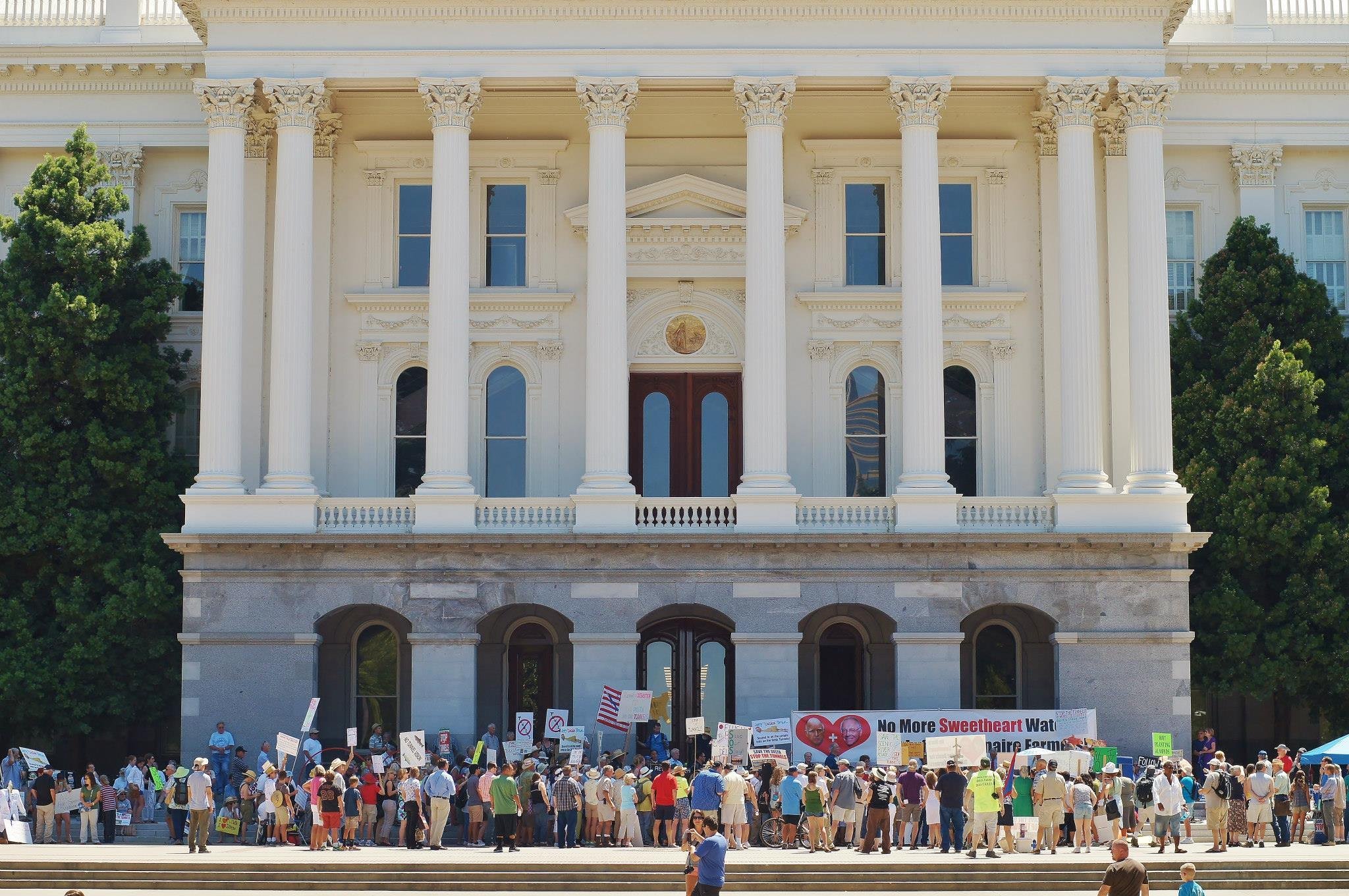 Protestors gathered in front of a large, white, neoclassical building with tall columns, holding signs and banners.