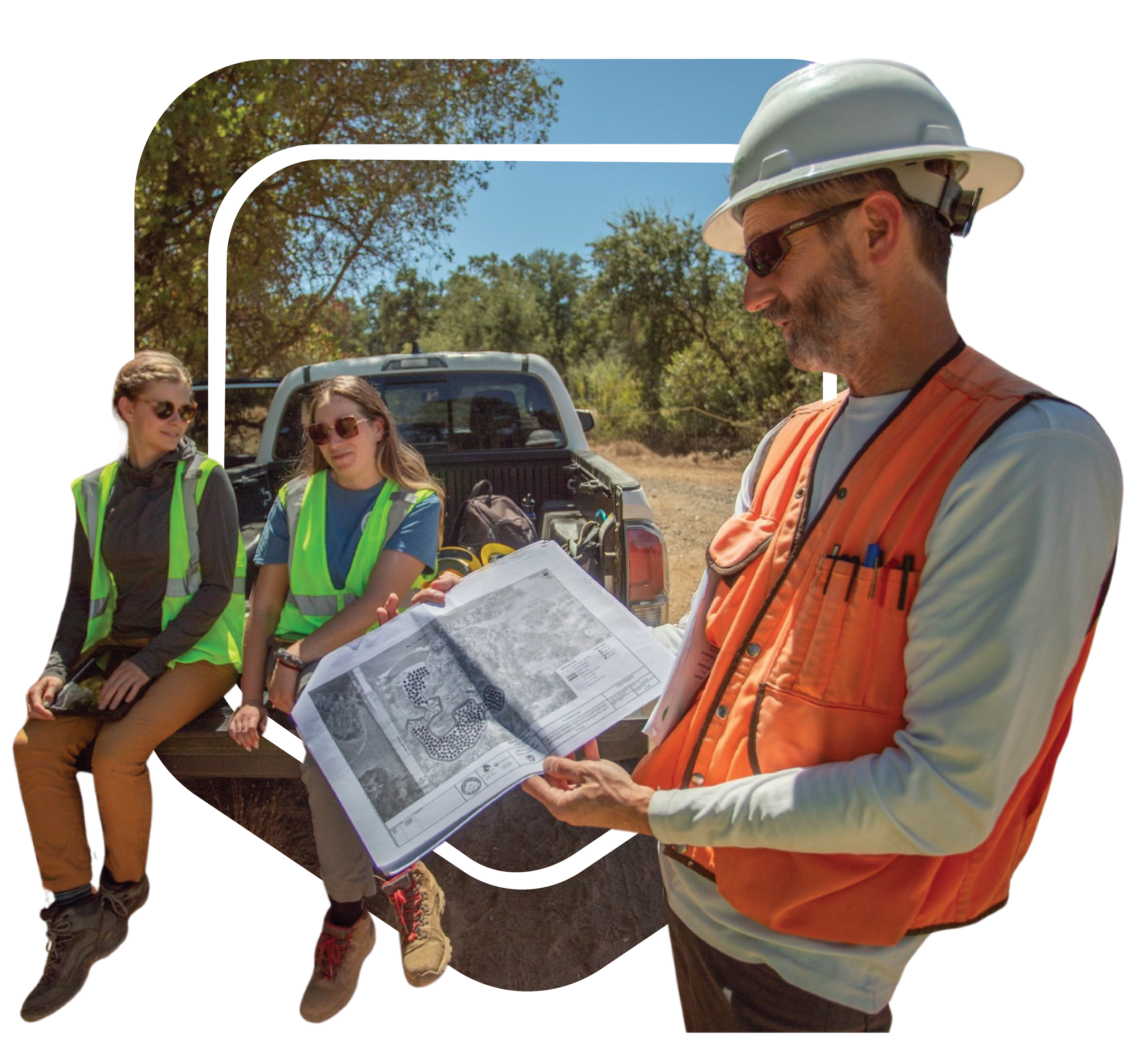 A man in safety gear holds a map while talking with two women in safety vests sitting on a pickup truck in a wooded outdoor area.