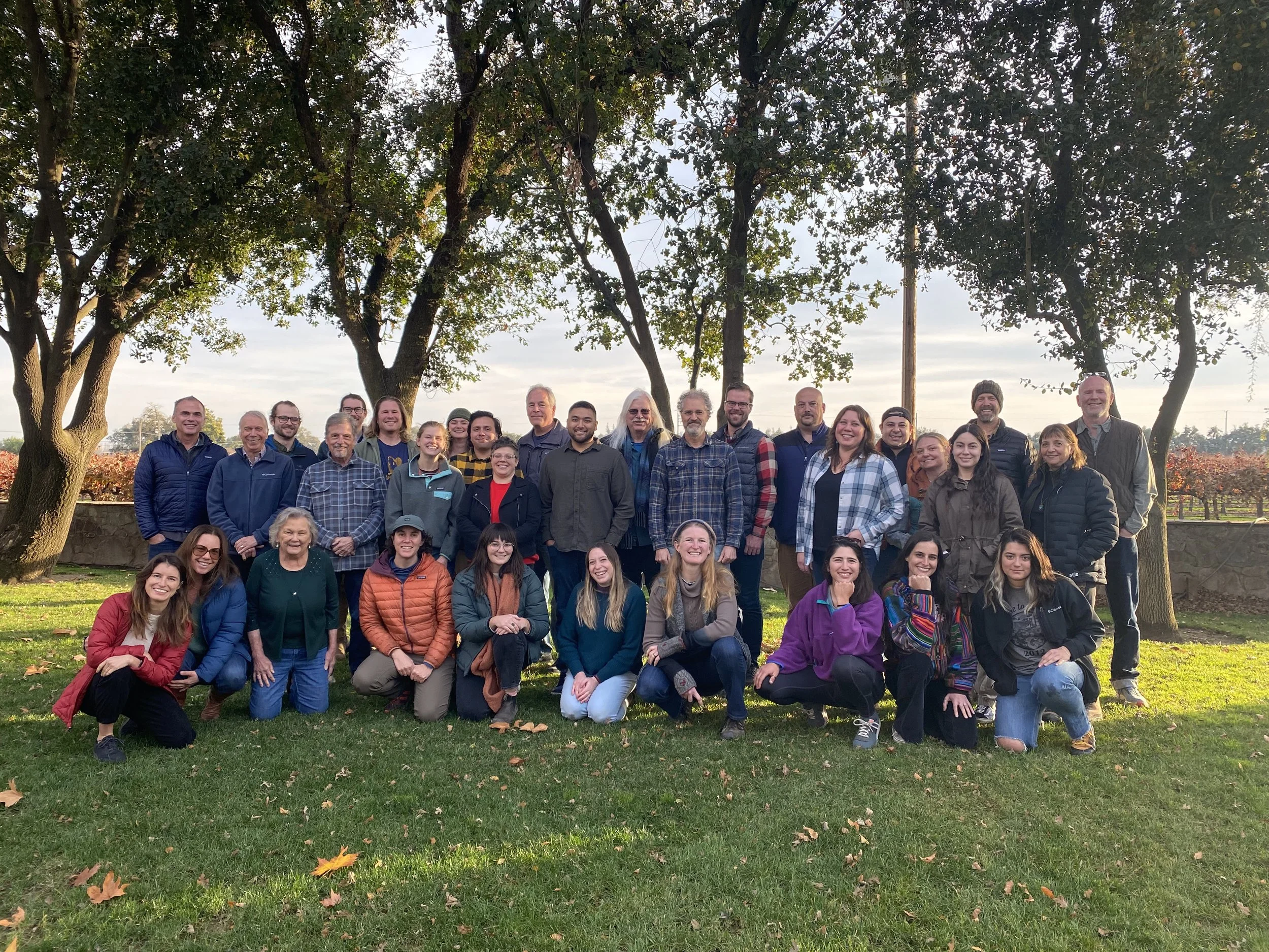 A large group of about 30 people outdoors on grass, posing for a group photo under trees with a clear sky in the background.