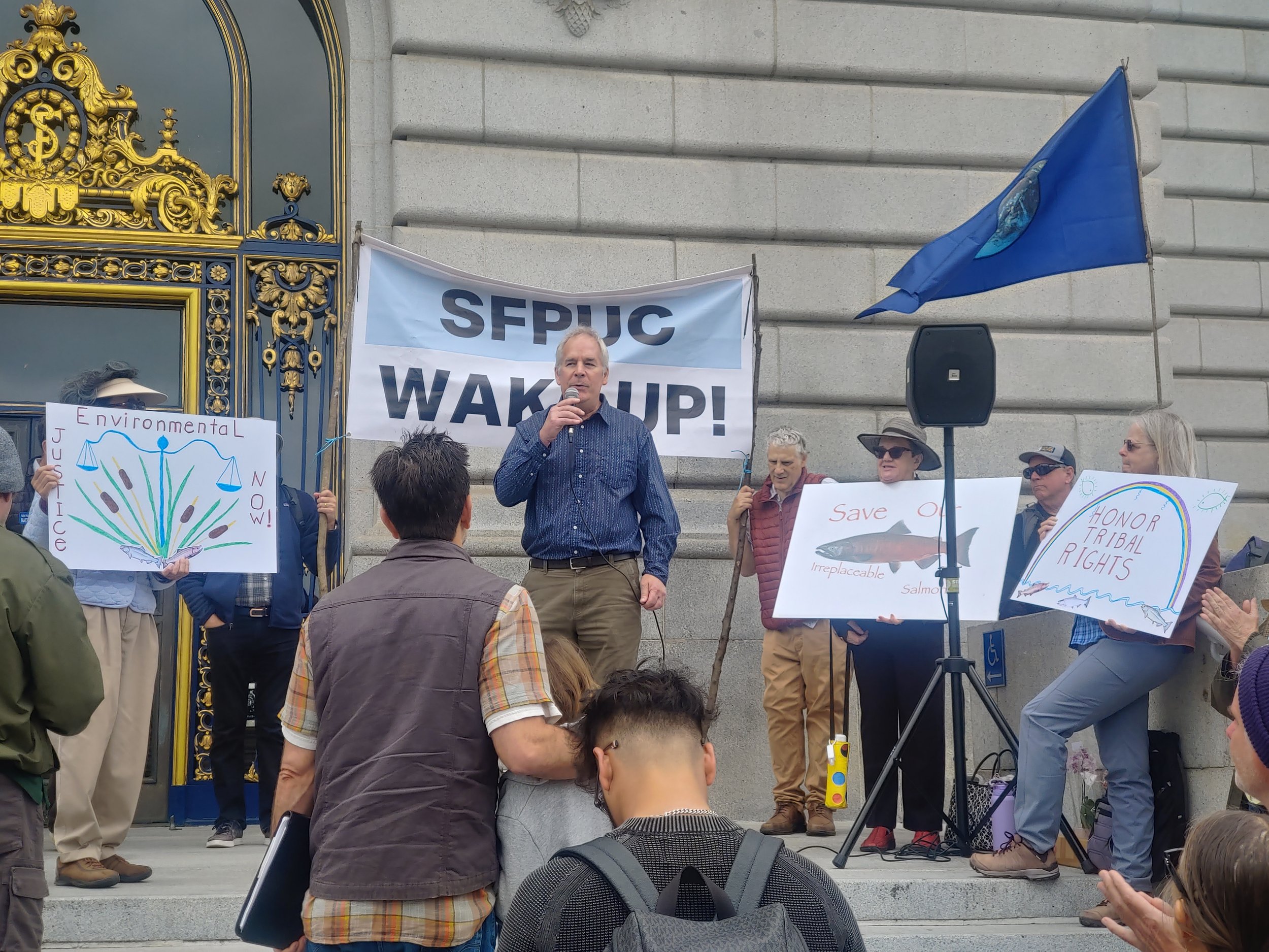 A group of people standing on the steps of a government building participating in a protest, holding signs advocating for environmental protection and tribal rights, with a man speaking into a microphone in the center.