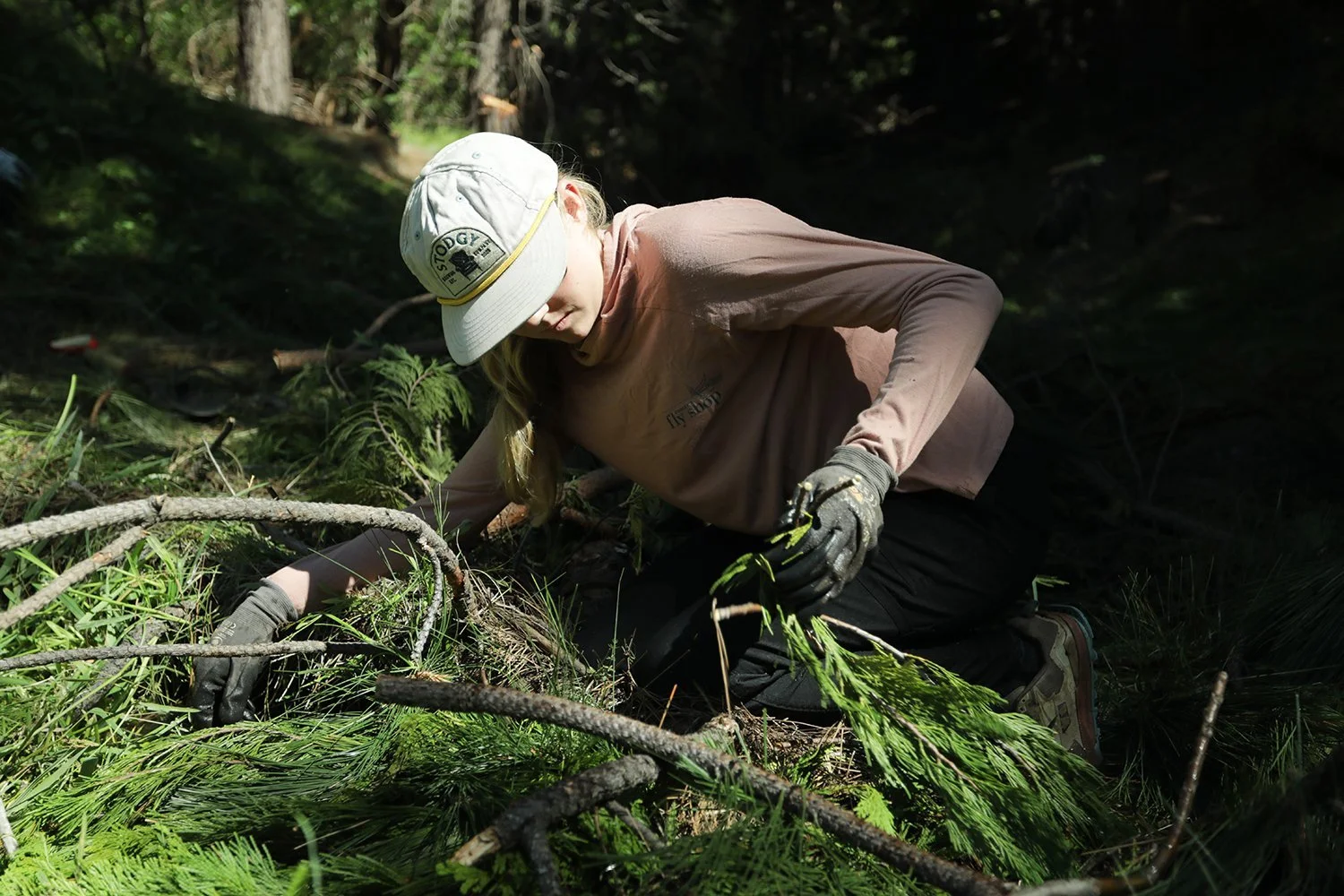 A person wearing a bucket hat, brown long-sleeve shirt, and gloves, crouching and removing debris from a forest floor covered with branches and green foliage.