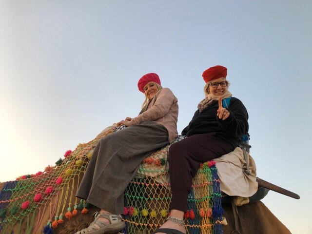 Two women sitting on a decorated camel, wearing red berets, with a clear blue sky in the background.