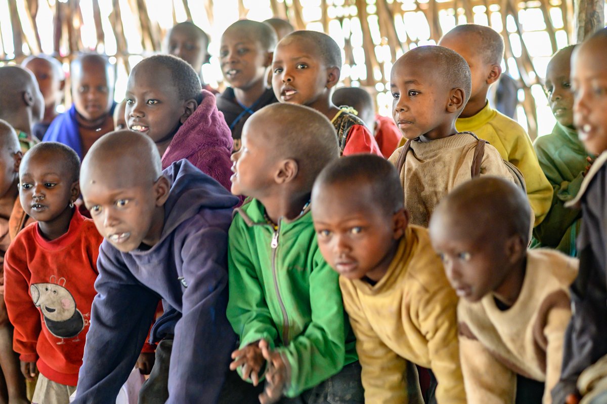 A group of children gathered indoors, some leaning forward, observing something attentively. They are dressed in colorful clothing, with a backdrop of wooden walls.
