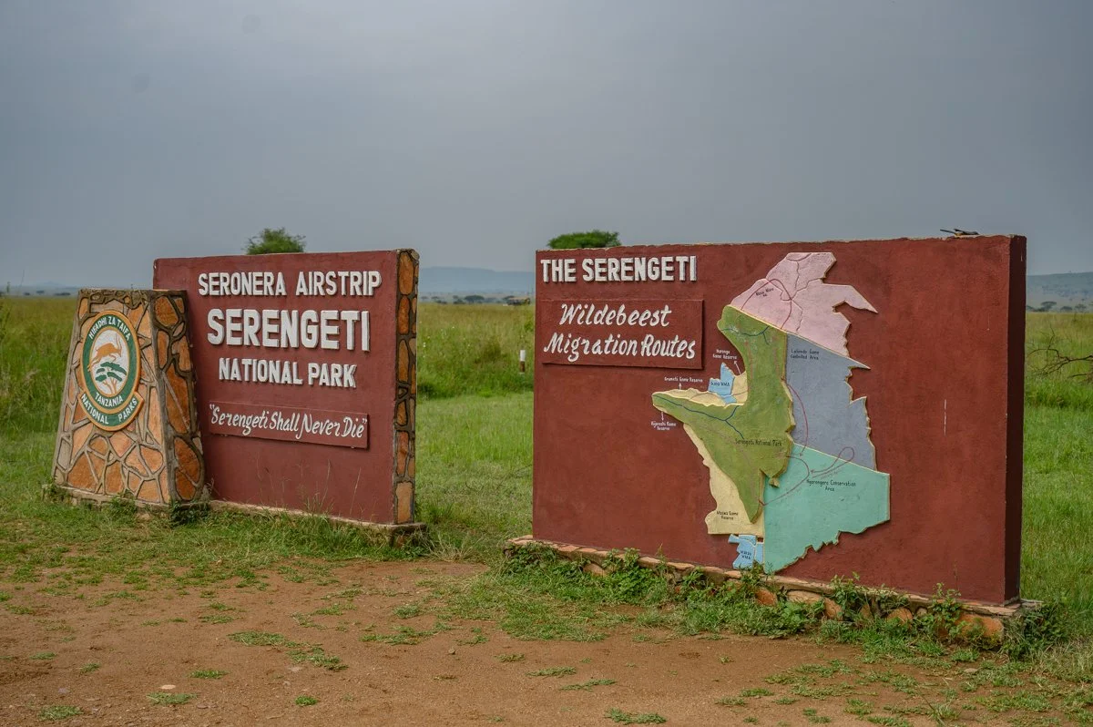Signpost at Serengeti National Park in Tanzania displaying maps of the Serengeti, with the slogan 'Serengeti Shall Never Die' and an emblem of the Tanzania National Parks, against a grassy landscape and cloudy sky.