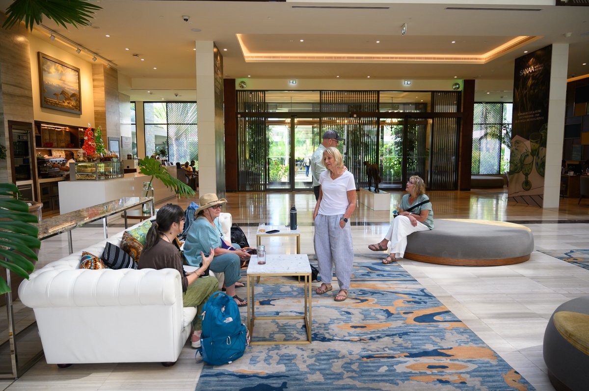 A hotel lobby with several people sitting and standing, relaxing and talking. There is a white sofa with three women seated, one in a hat, another in a brown shirt, and the third in a green shirt. A woman in white pants is sitting on a large gray rou