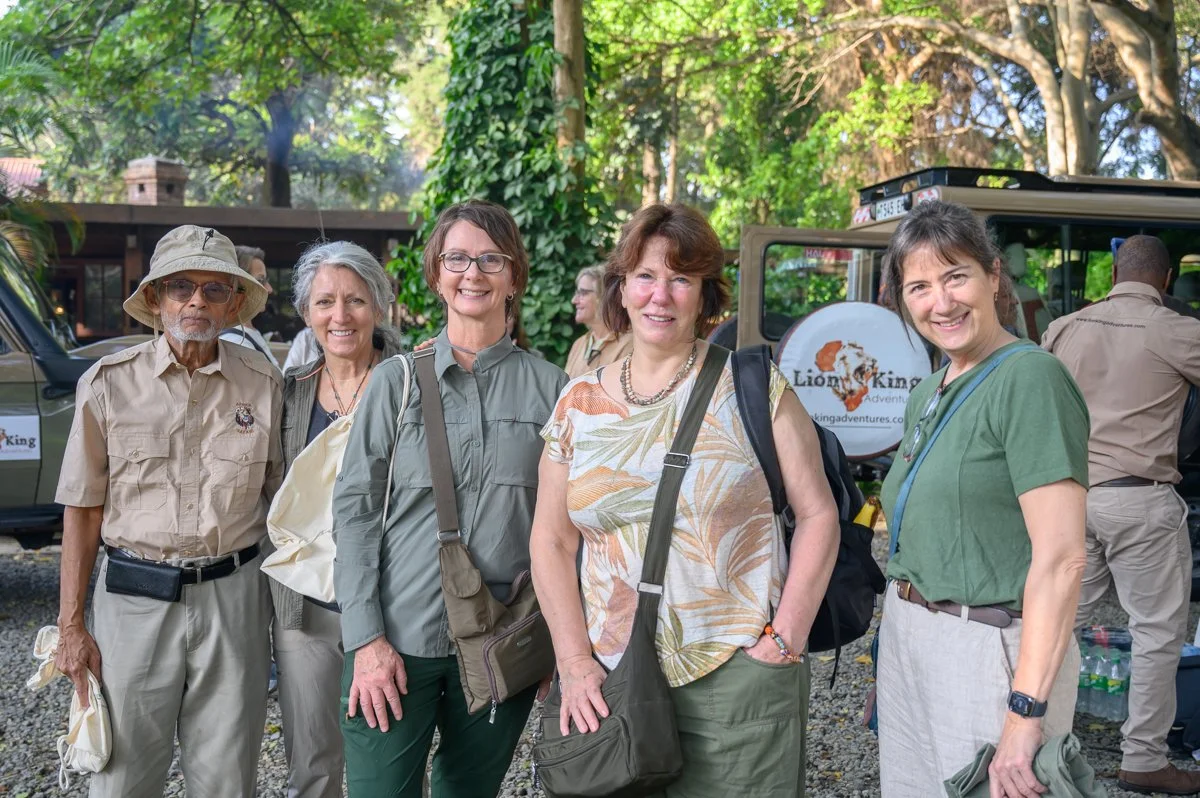 A group of five women and one man standing outdoors in a jungle or forest setting, posing for a photo with a lion king adventure sign in the background, some with backpacks and wearing casual outdoor clothing.