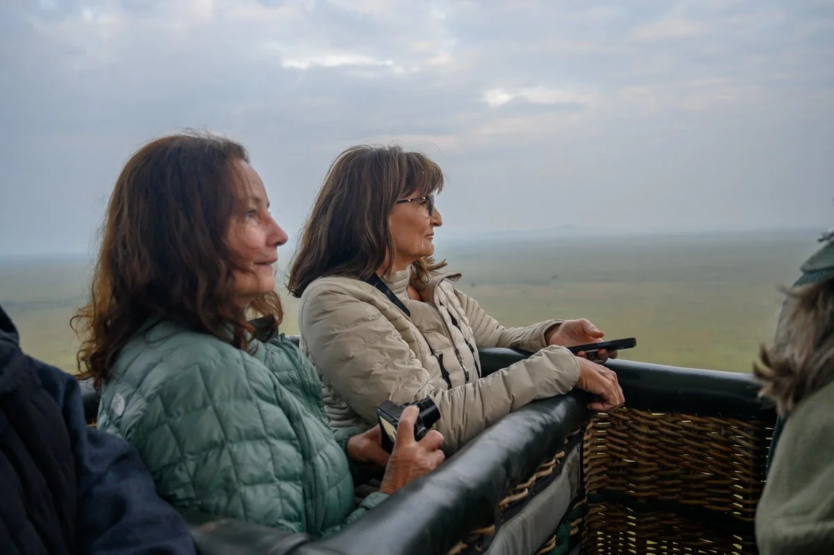 Two women in a hot air balloon basket overlooking a flat landscape, with cloudy skies above.