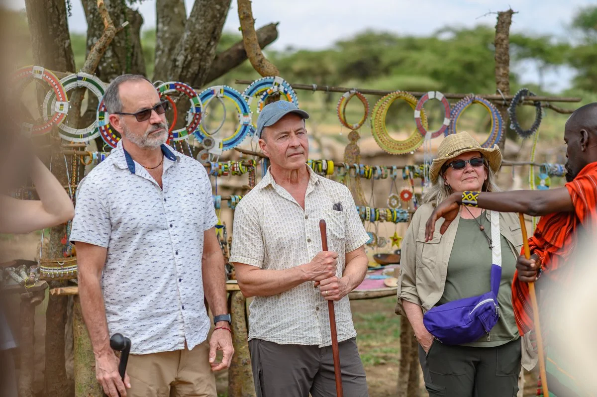 Three tourists, two men and one woman, are at a market stall selling colorful beaded jewelry in an outdoor setting with trees. They are attentively listening to a local man explaining something about the jewelry.