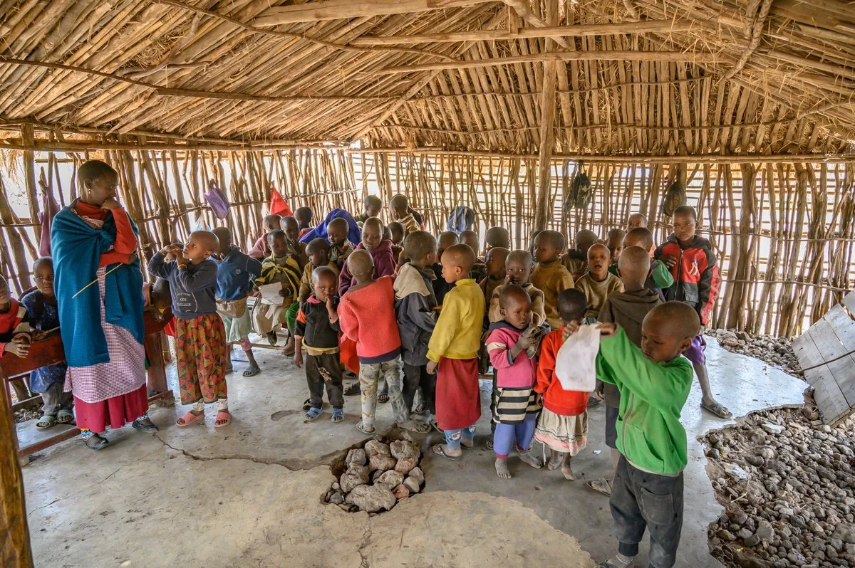 A group of children inside a makeshift classroom with a woman standing among them, under a roof made of wooden sticks and thatched material.