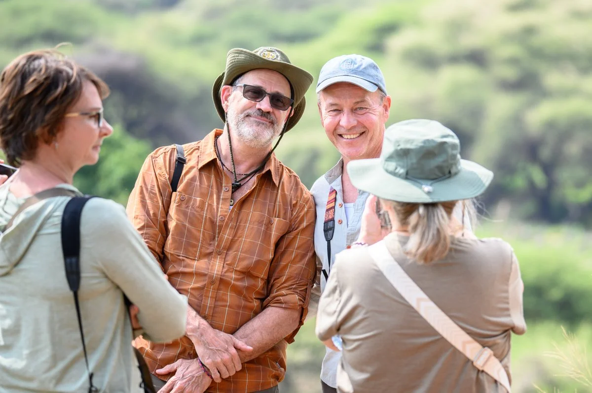 Five people outdoors smiling and talking together in a natural setting, dressed in casual hiking clothes and hats.