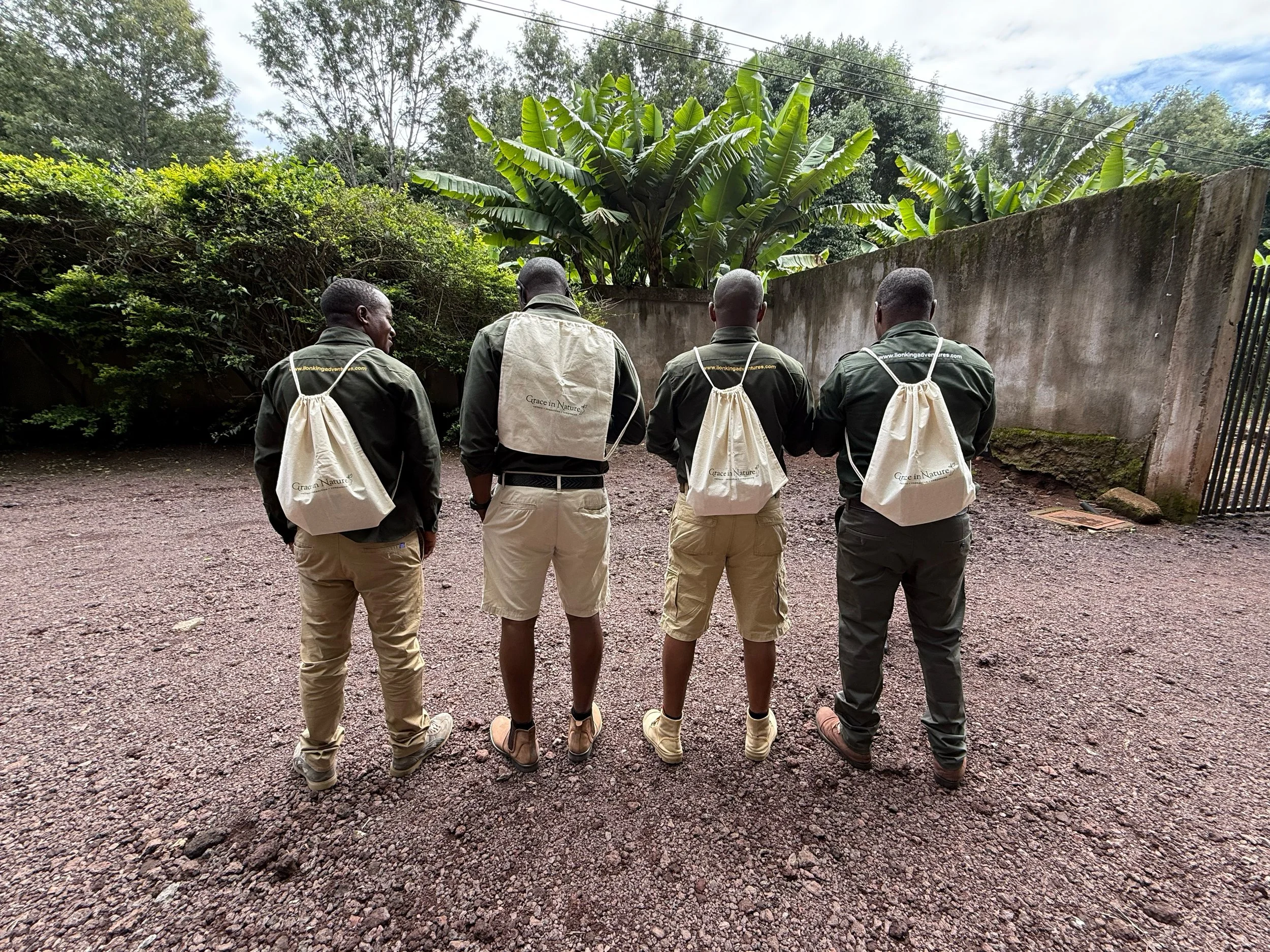 Four men standing outdoors, facing away, wearing matching beige backpacks and similar dark clothing, with lush green trees and a concrete wall in the background.