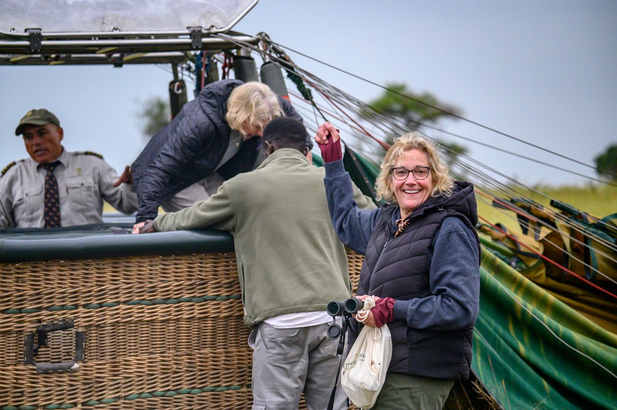 Smiling woman with glasses and curly blonde hair holding binoculars, standing next to a hot air balloon basket, with a group of people preparing the hot air balloon for flight.