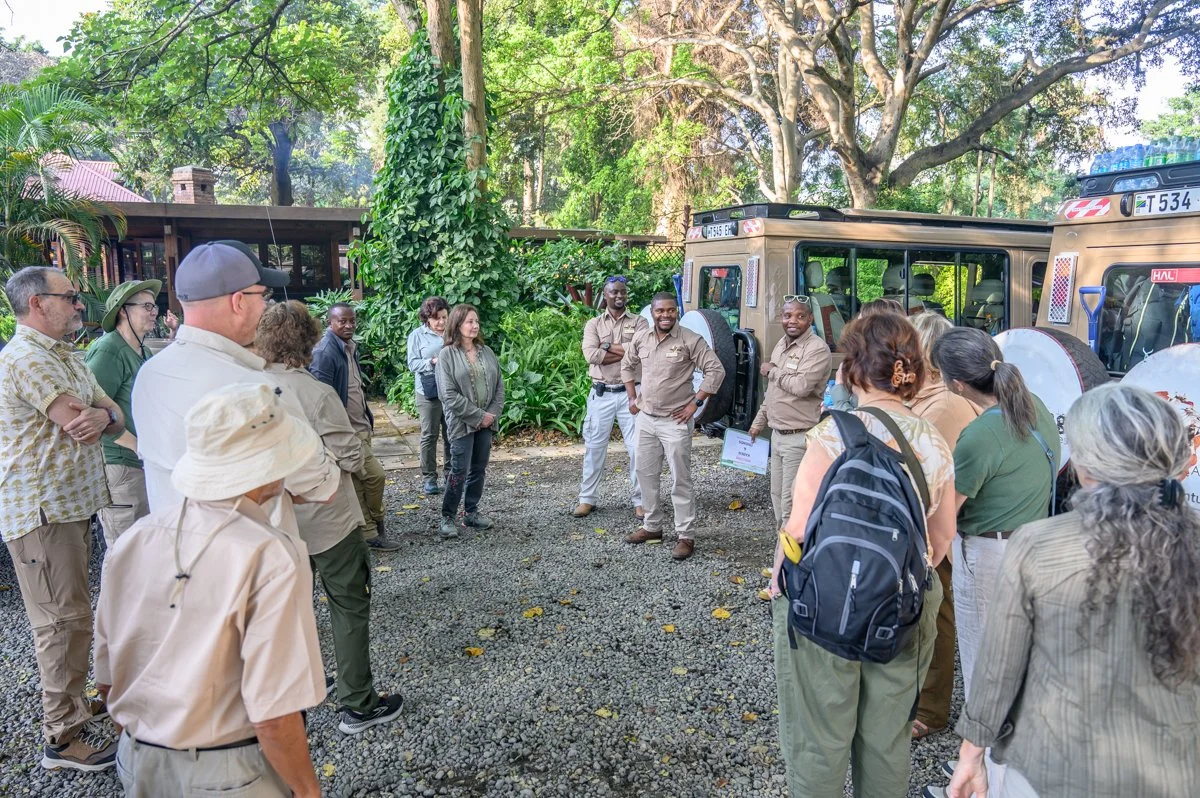 Group of people listening to guides outside near safari vehicles, surrounded by trees and greenery.