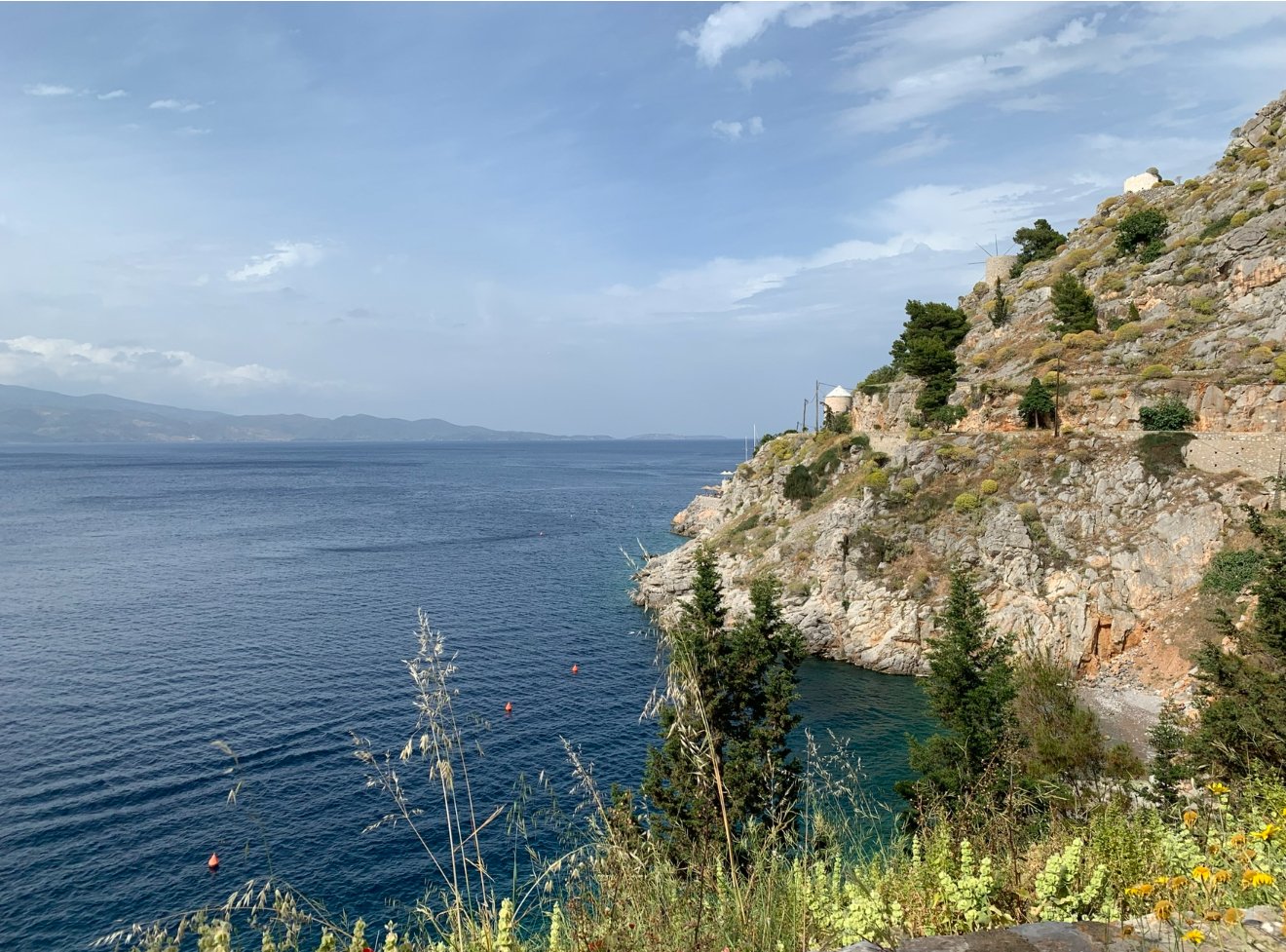 A coastal landscape with blue water, rocky cliffs covered in greenery, and a partly cloudy sky.