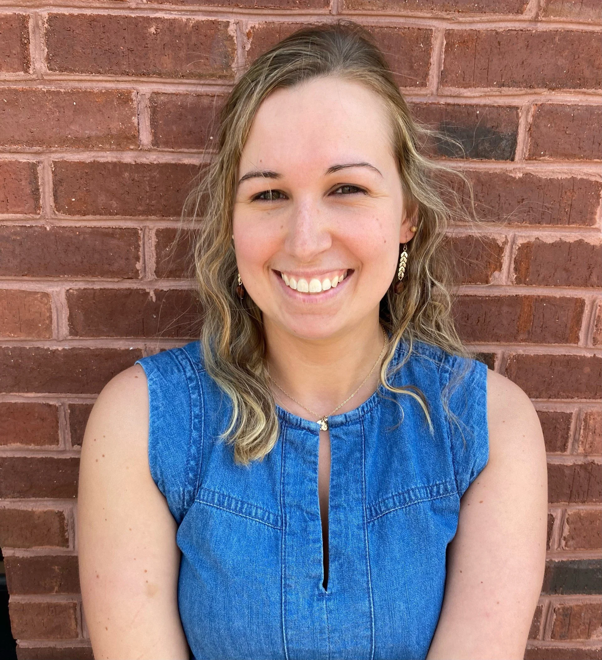 A woman with curly blonde hair smiling, wearing a sleeveless blue denim top, standing in front of a red brick wall.
