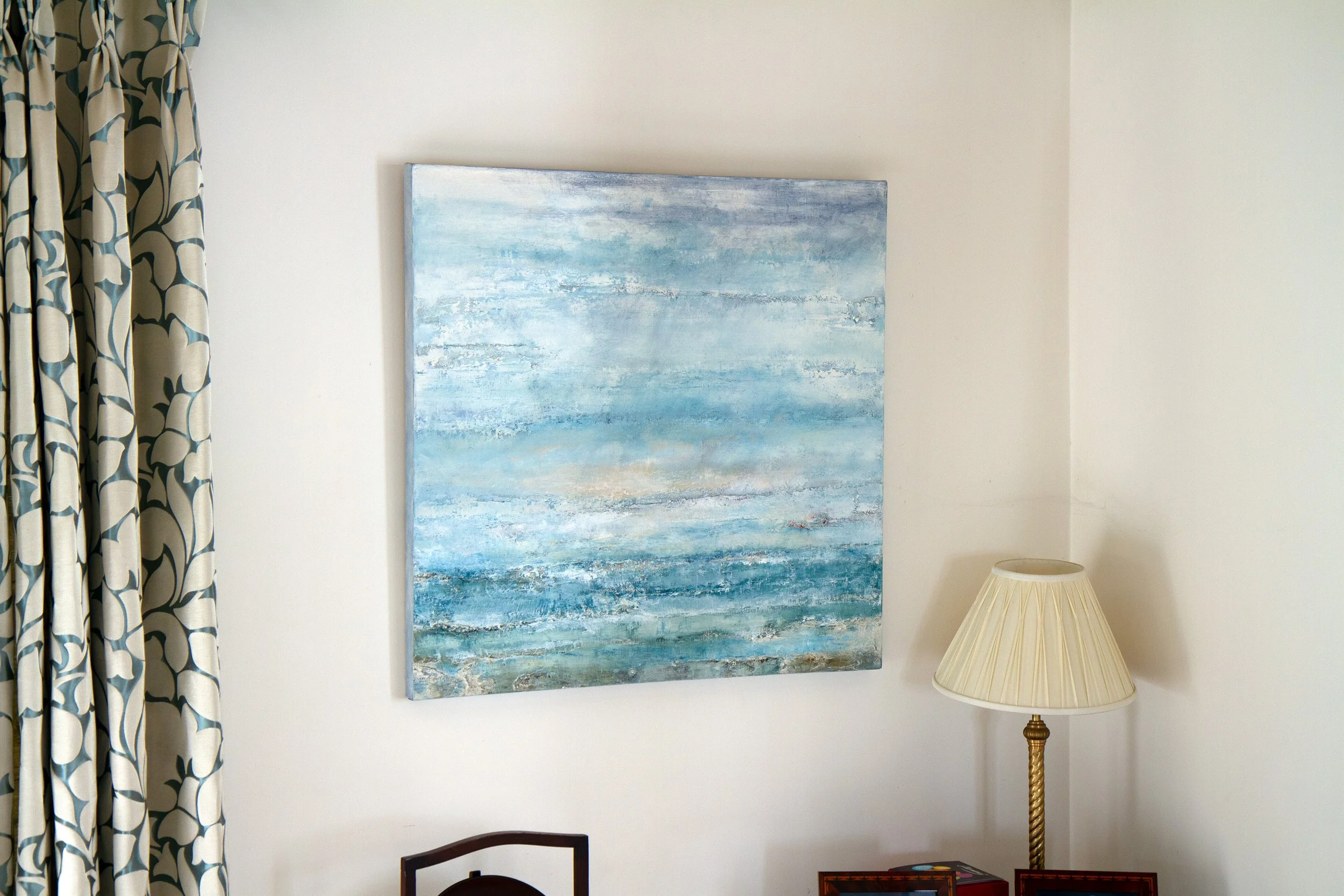 A room corner with a large abstract painting of blue tones on the wall, a beige and blue patterned curtain on the left, a white/cream table lamp with a gold stand on the right, and part of a dark wooden chair at the bottom.