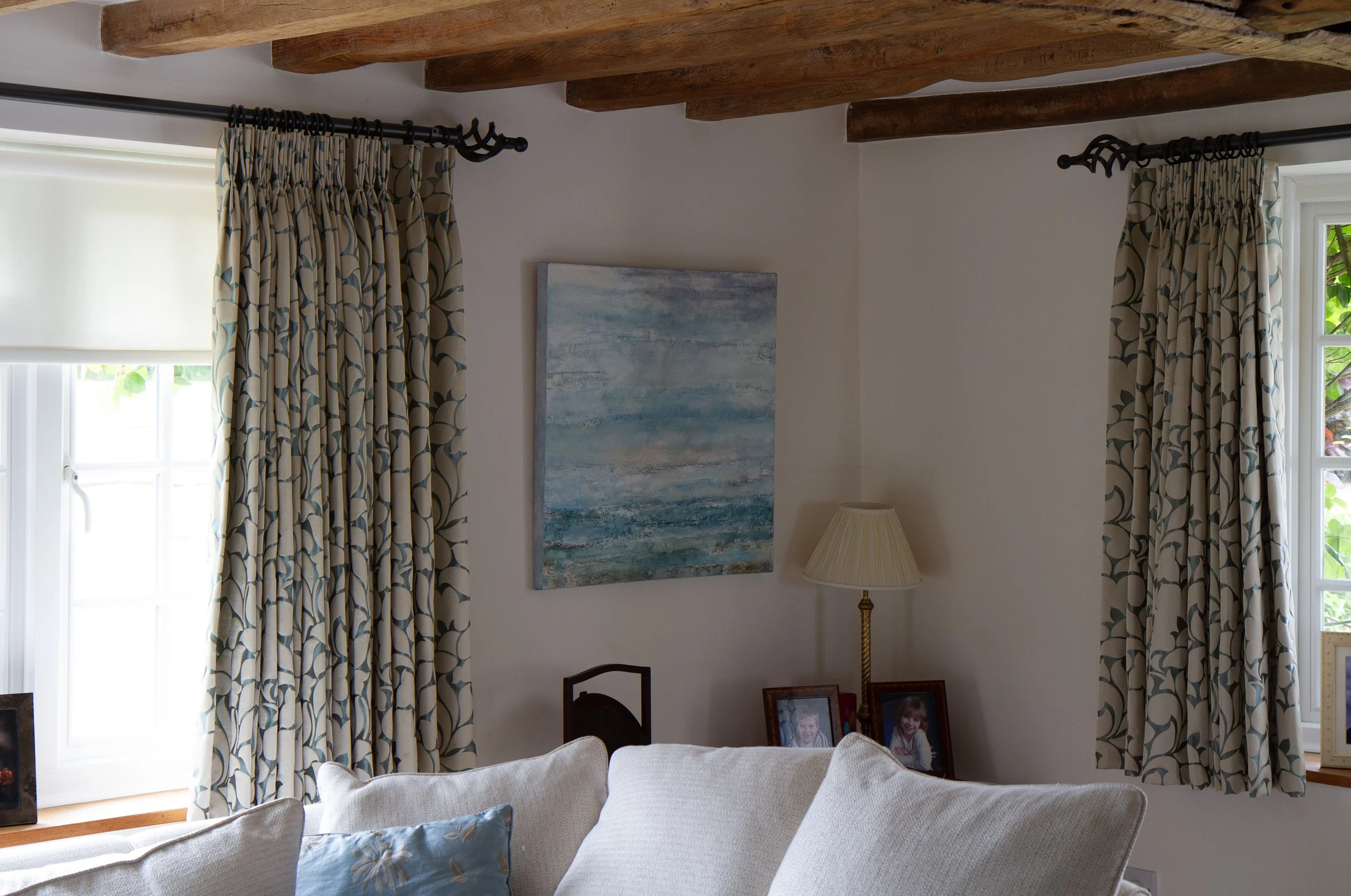 A cozy living room with cream-colored sofa and a blue floral pillow, two windows with patterned curtains, a painting on the wall, a lamp with a beige shade, and family photographs on a side table.