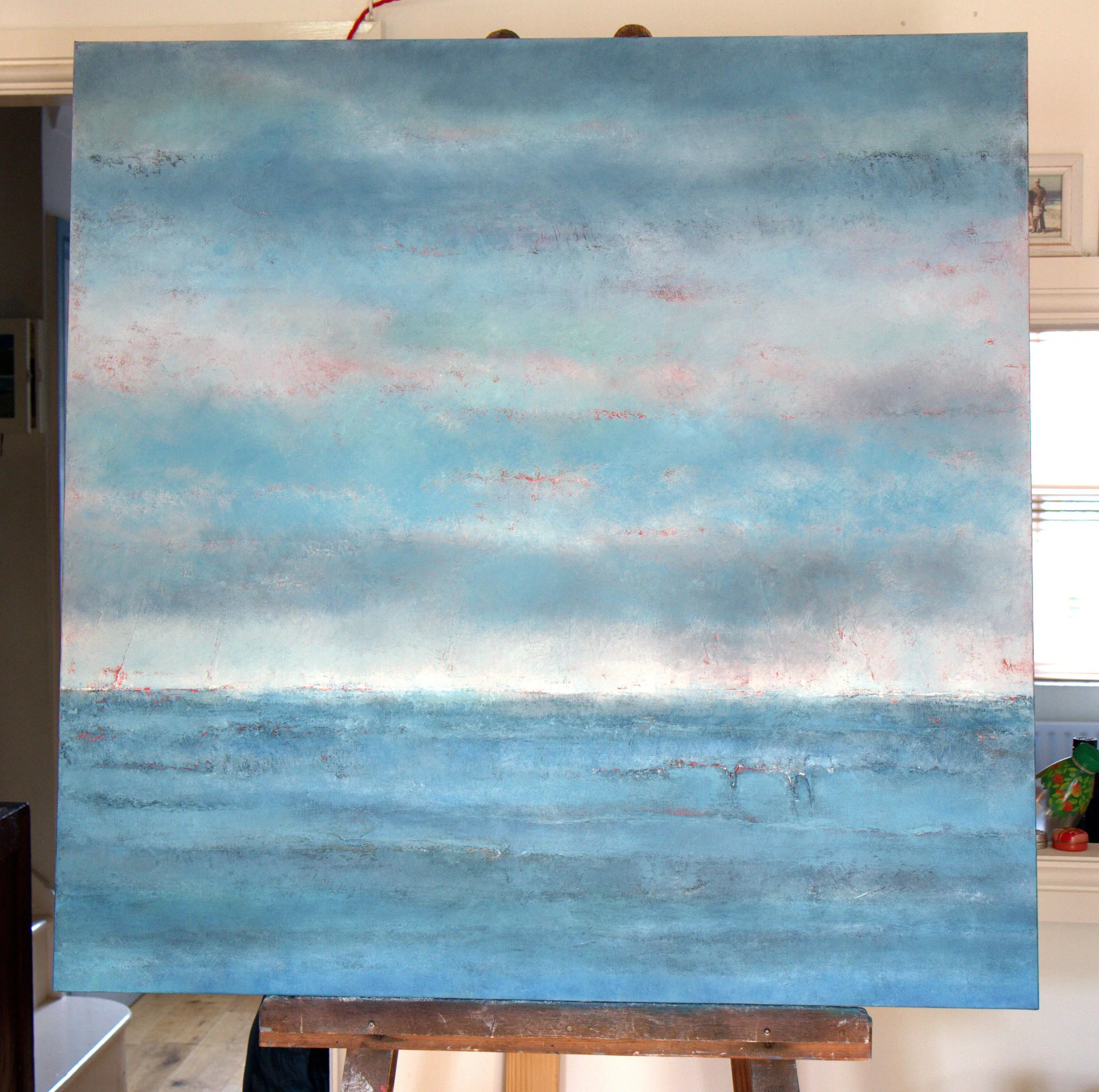 A large abstract landscape painting on a blue sky and ocean with pink highlights, hung on an easel in an indoor room.