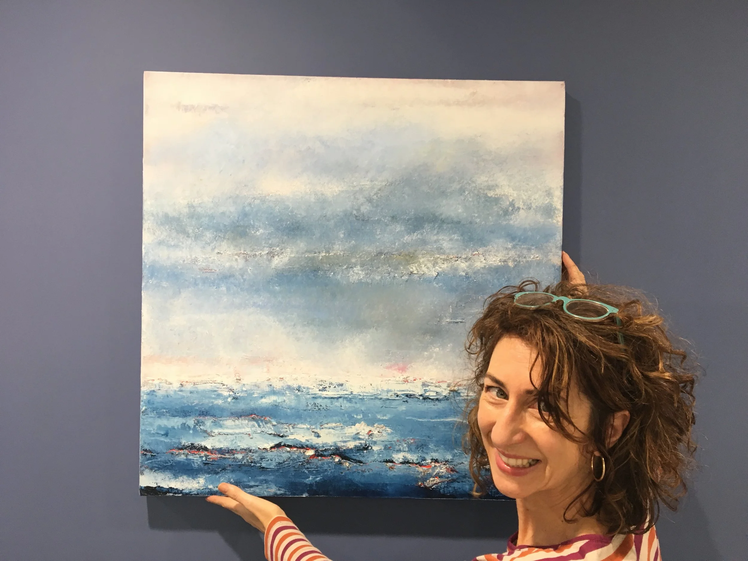 A woman holding up a large seascape painting with blue and white hues, standing in front of a plain dark blue wall.