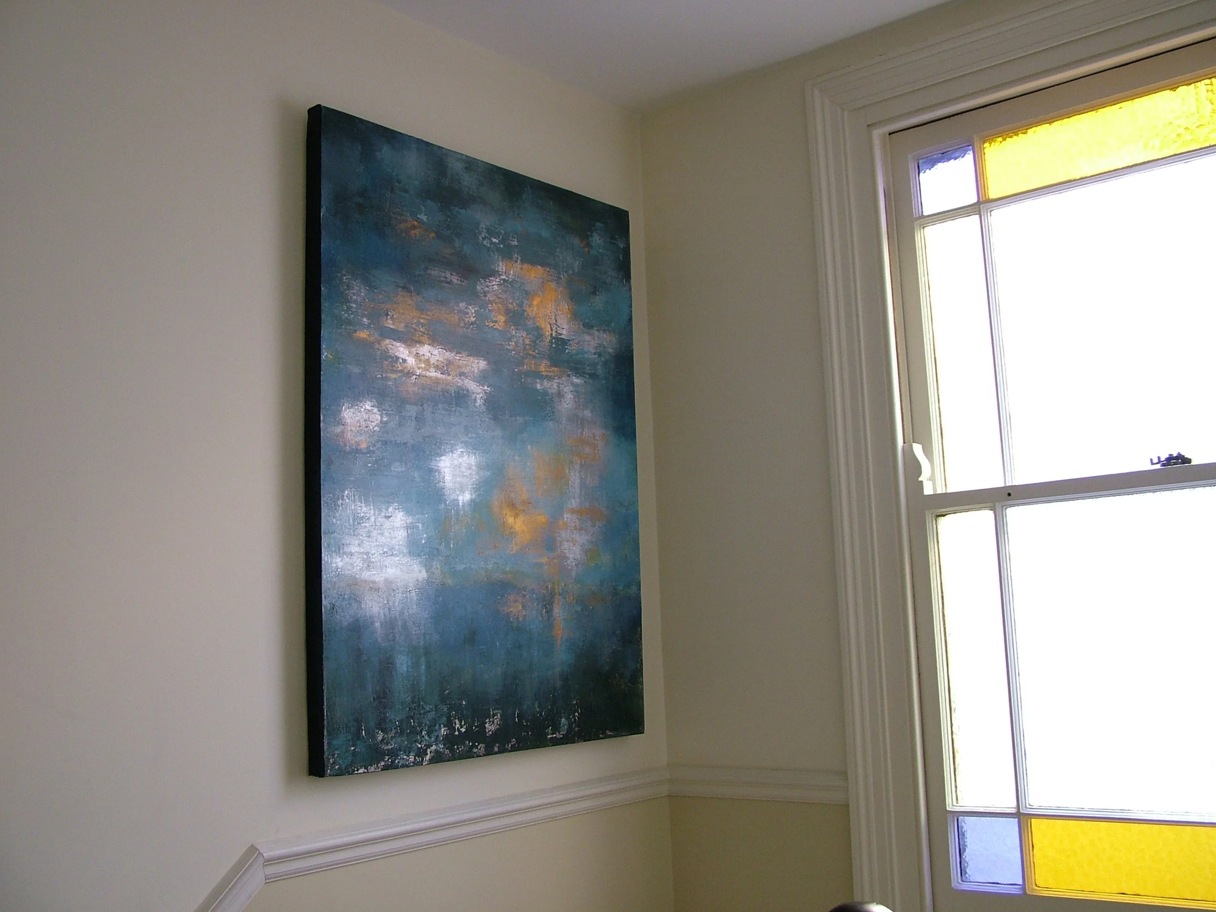 An abstract painting with shades of blue, white, and hints of gold hanging on a white wall in a room with a window that has white trim and colorful stained glass panels.