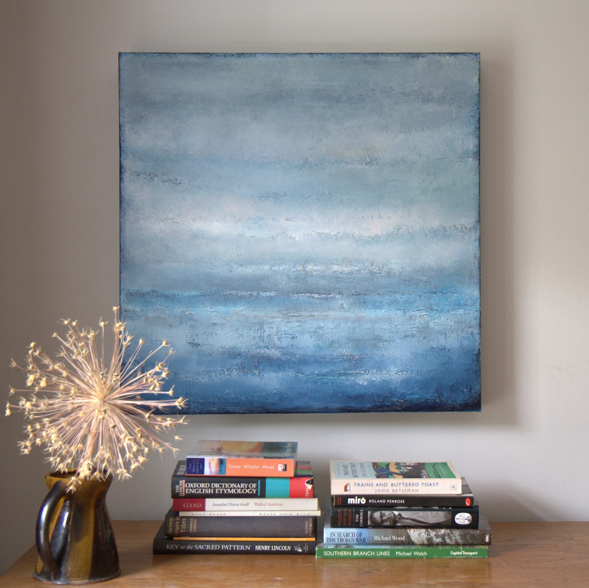 A table with a stack of books, a decorative lamp, and a large blue abstract painting on the wall behind.