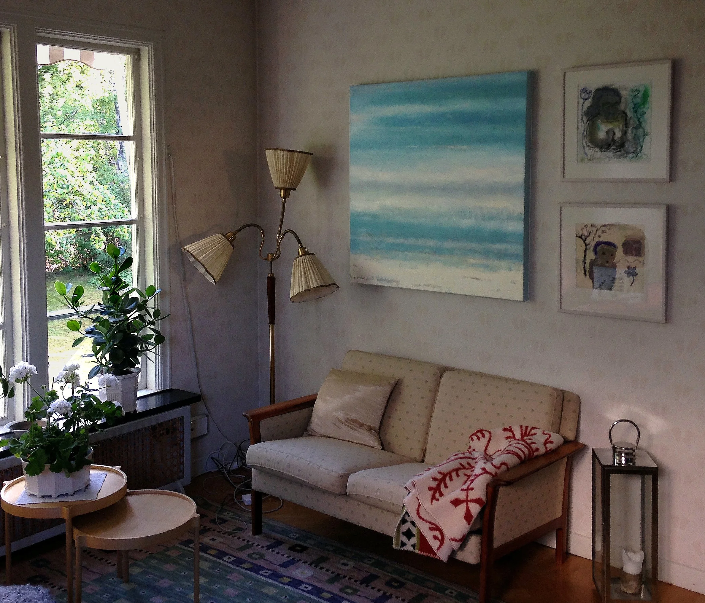 A cozy living room with a beige sofa, a small side table with a candle, a tall floor lamp, a window with green trees outside, houseplants on a table, and several framed artworks on the wall.