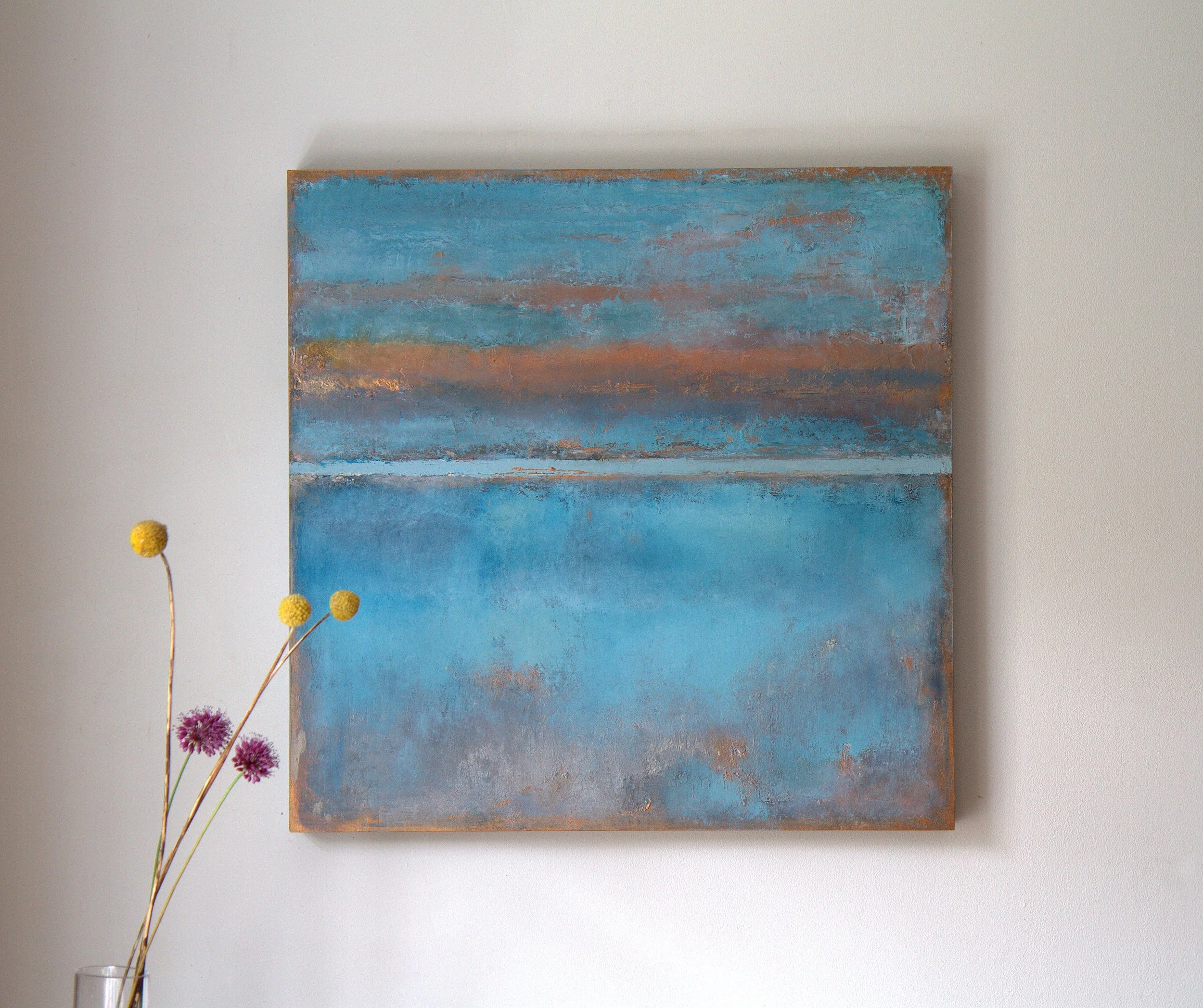 Abstract painting with blue, orange, and gray tones hanging on a light-colored wall. A glass vase with yellow and purple flowers is in front of the painting.