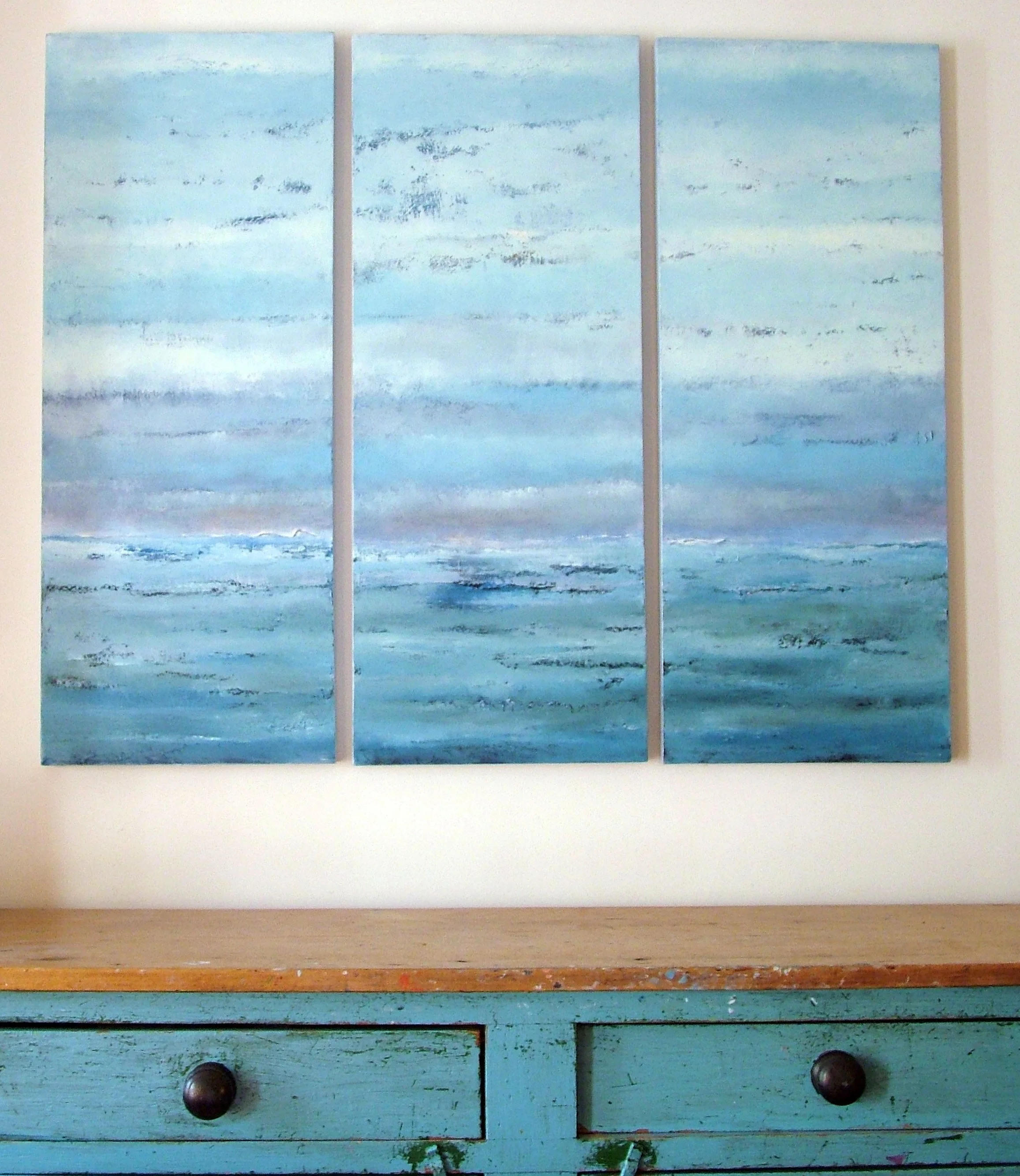 Abstract painting of a seascape with shades of blue, white, and gray, divided into three panels, hanging above a distressed turquoise wooden dresser.