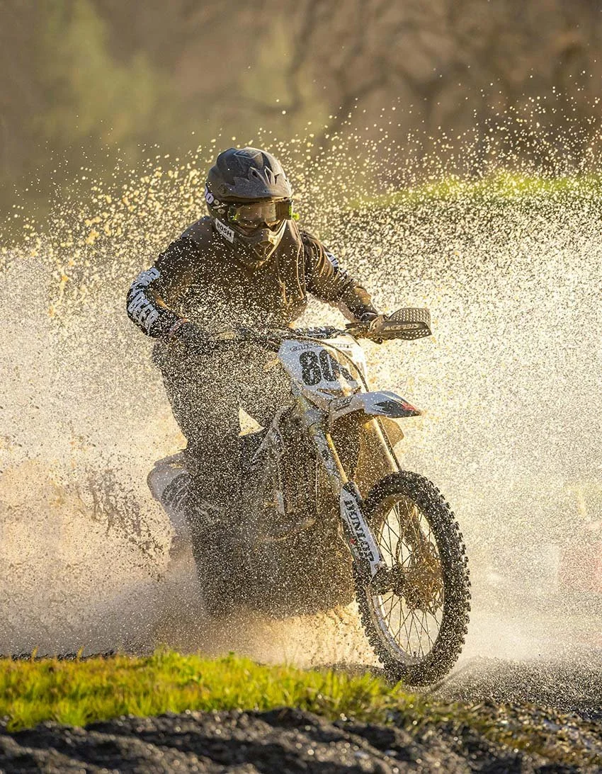 A person riding a dirt bike through water, creating a splash, wearing a black helmet and protective gear.