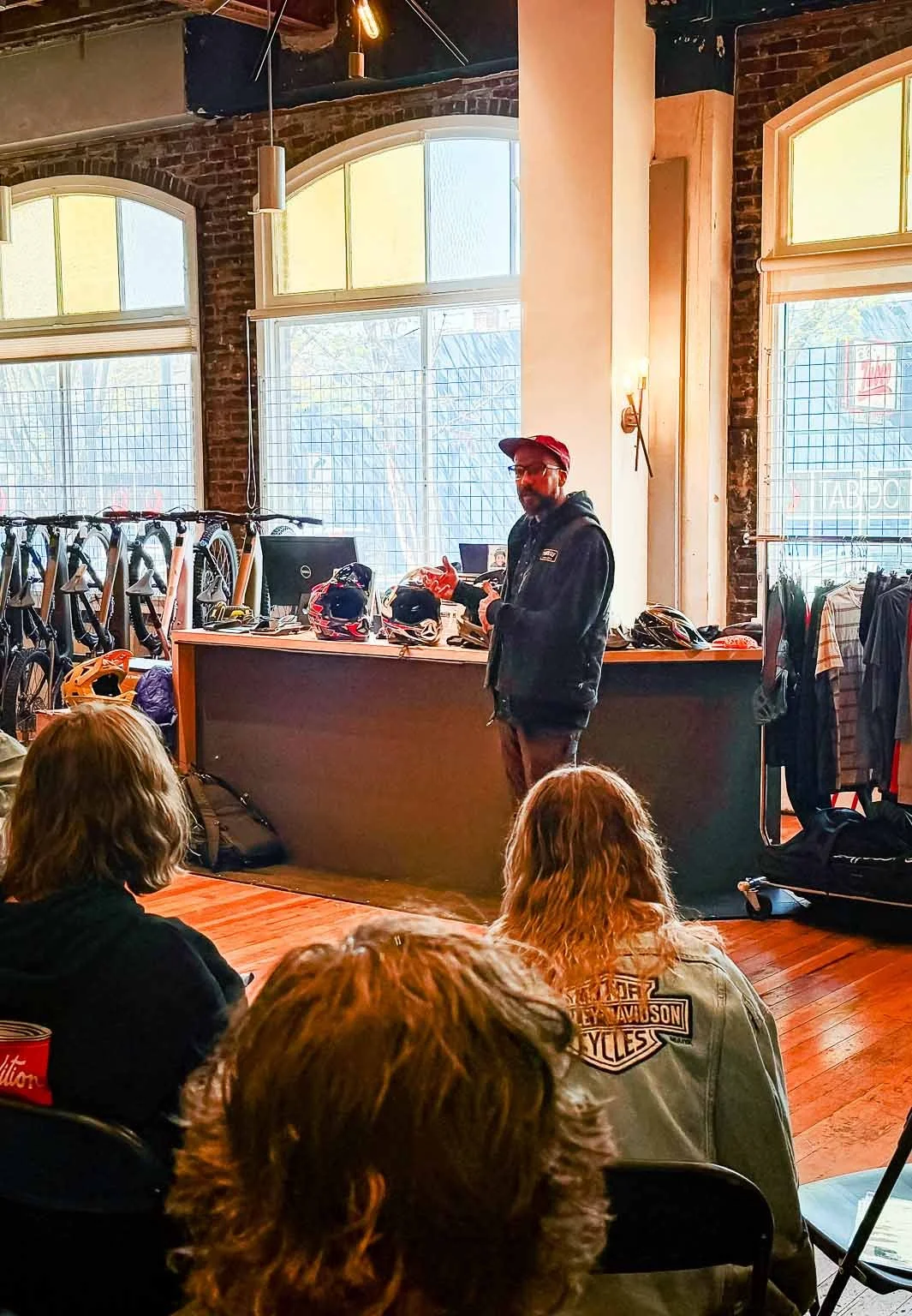 A man in a hoodie and baseball cap standing behind a counter with helmets, addressing a group of people seated in front of him in a bike shop with large windows and bicycles on display.