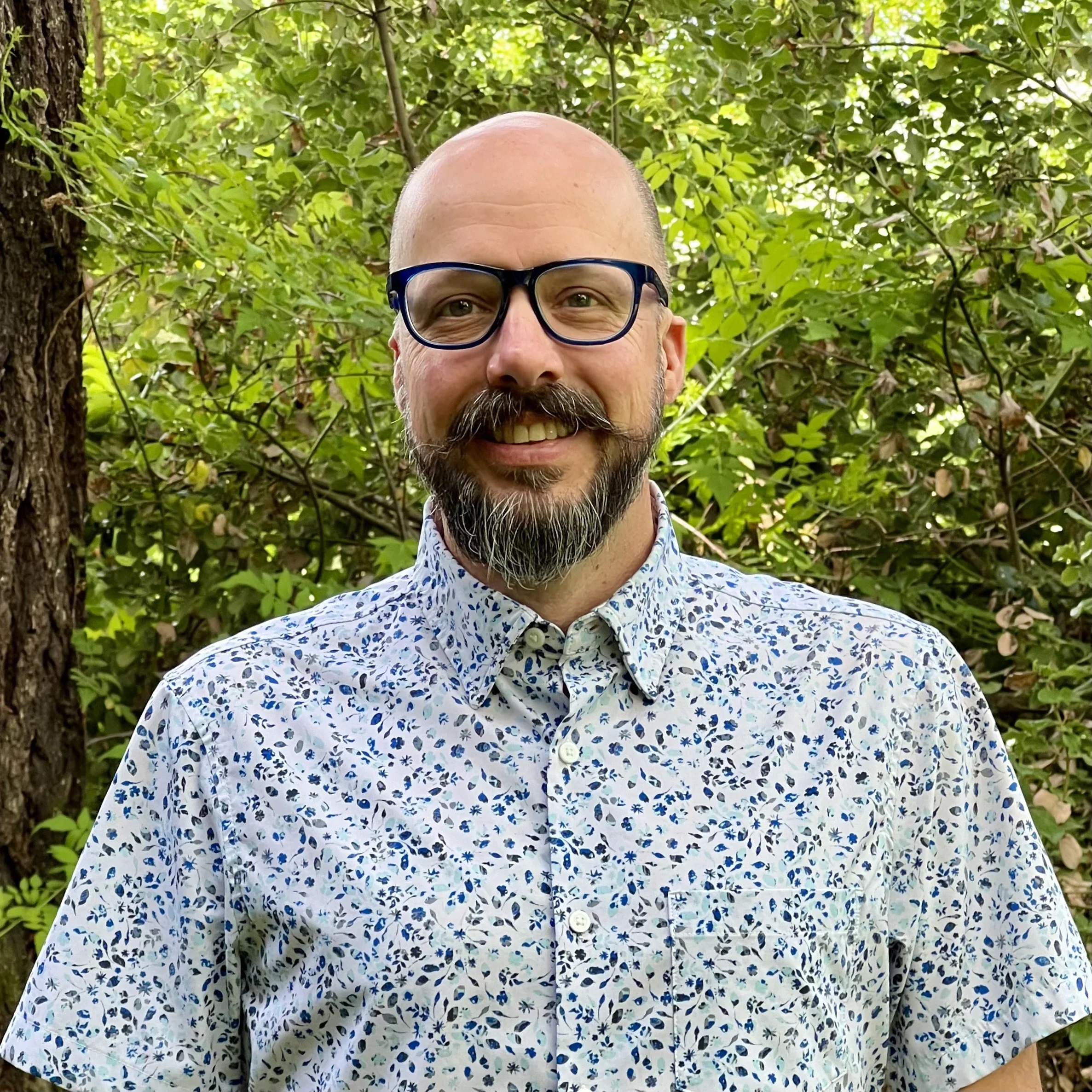 A smiling man with glasses, a beard, and a mustache wearing a light-colored shirt with a blue floral pattern, standing outdoors among green trees and foliage.