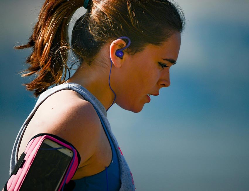 A woman running outdoors, wearing earphones with a smartphone attached to her arm.