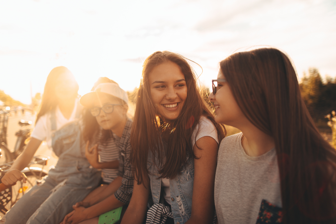 Four girls sitting on a bench outside during sunset, smiling and talking, with bicycles in the background.