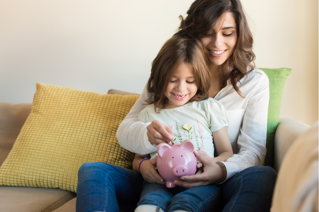 Mother and daughter sitting on a sofa, smiling, with the daughter holding a pink piggy bank.