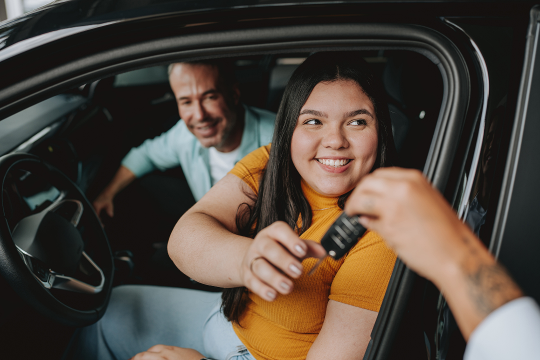 A young woman receiving car keys after buying a car while sitting in the driver's seat, with a man sitting in the passenger seat.