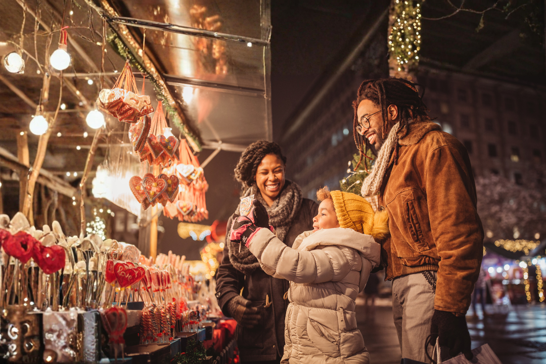 A smiling family shopping at an outdoor holiday market at night, with decorated stalls and Christmas lights.