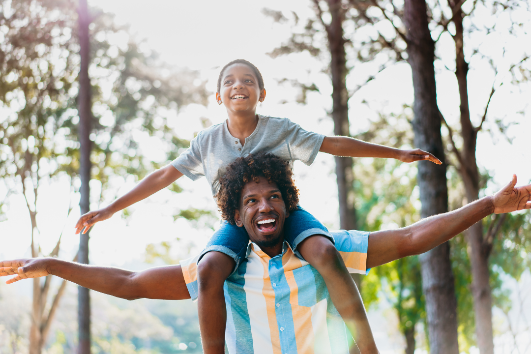 A man giving a piggyback ride to a smiling young girl with arms outstretched, outdoors in a wooded area during daytime.