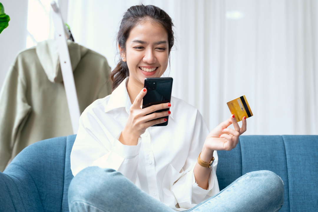 A young woman sitting on a blue couch, smiling and looking at her smartphone while holding a credit card in her other hand.