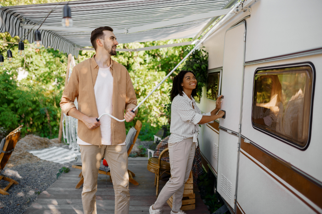 A young man is pulling down an awning attached to a camper trailer as a young woman unlocks the camper door, with outdoor furniture and greenery in the background.