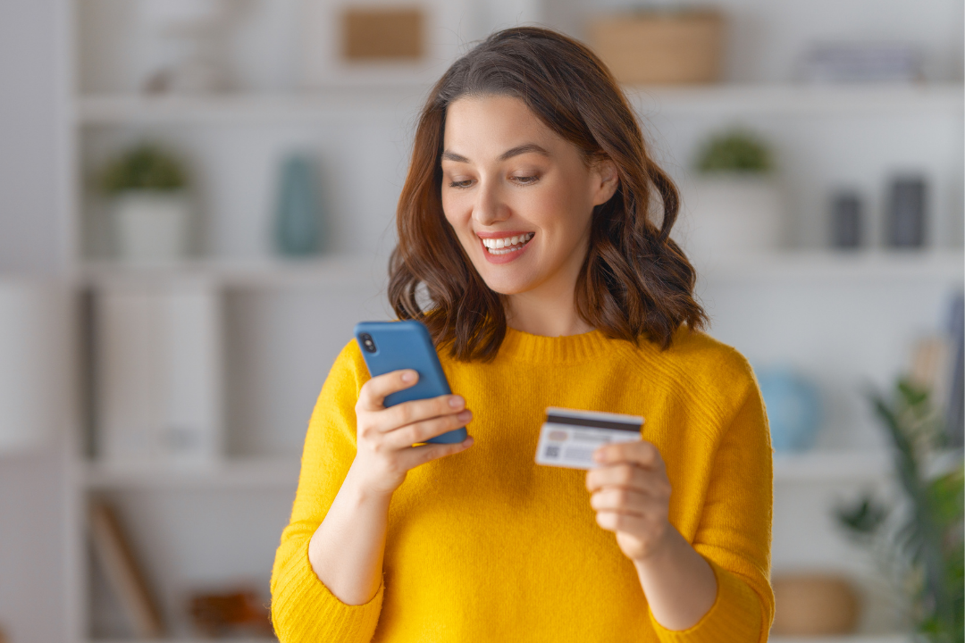 A woman with shoulder-length brown hair wearing a yellow sweater, smiling happily while holding a smartphone in one hand and a credit card in the other, standing in a modern, bright room.
